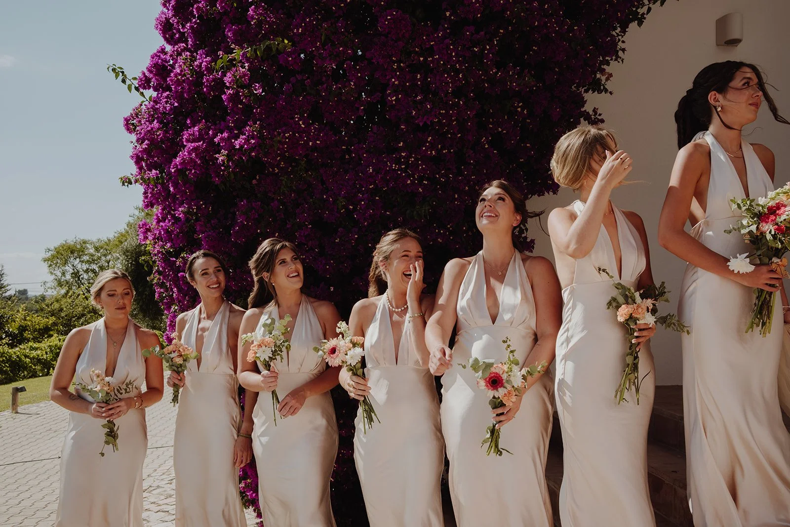 Group of women in white dresses holding bouquets, standing outdoors near a large purple flowering bush, smiling and laughing.