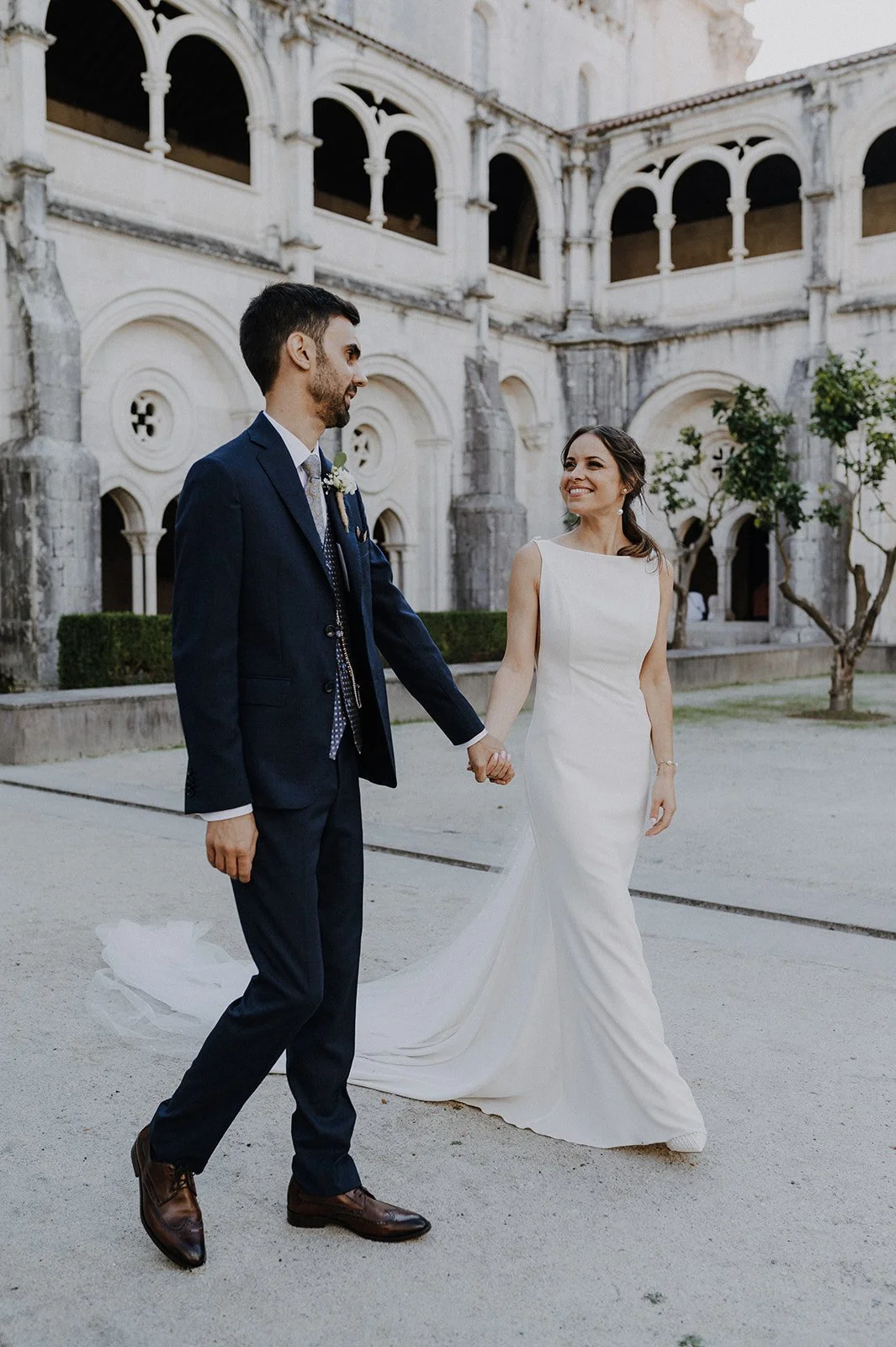 A bride and groom holding hands, smiling and walking outside near an old stone building with arches, during their wedding.
