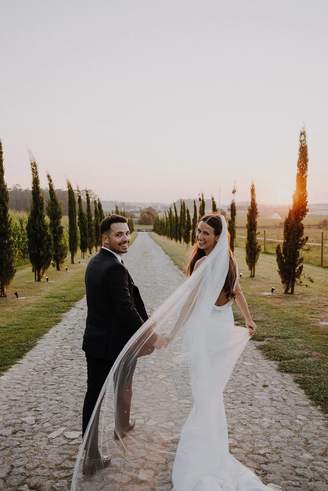 A newlywed couple holding hands and smiling at each other on a stone pathway lined with tall cypress trees at sunset.