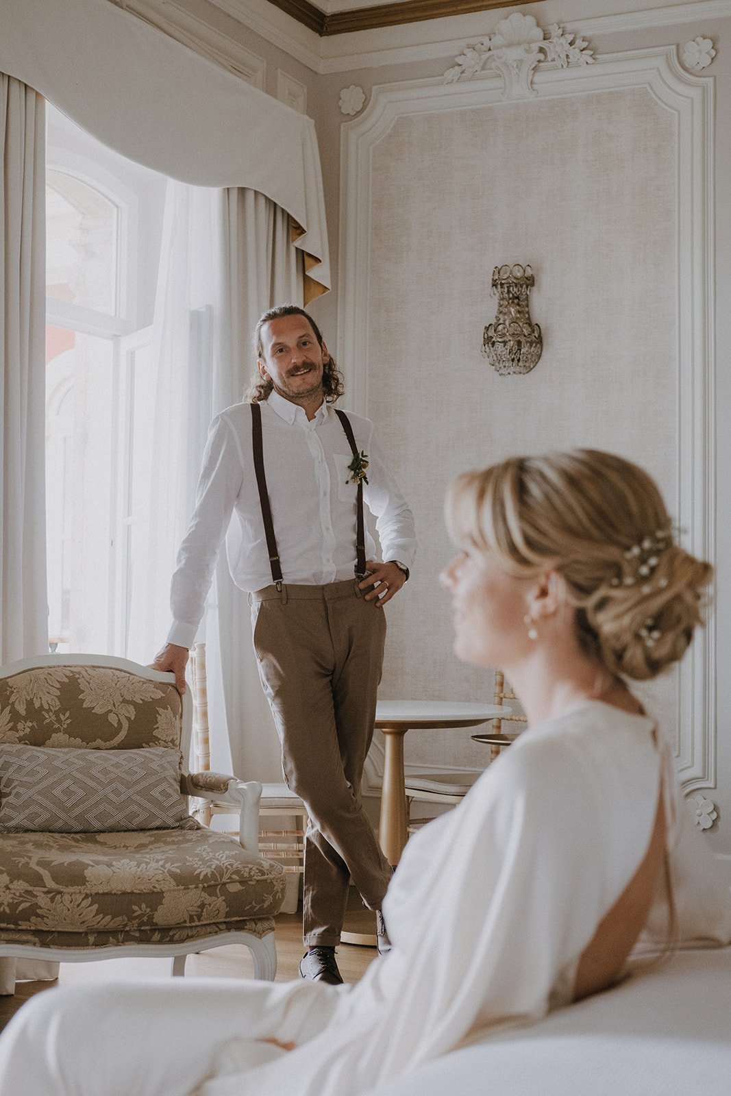 A man with long hair and a mustache, dressed in a white shirt with suspenders and tan pants, standing and leaning on a chair, smiling at a woman with blonde hair styled in an elegant updo, wearing a white dress, sitting on a sofa in a decorated room 