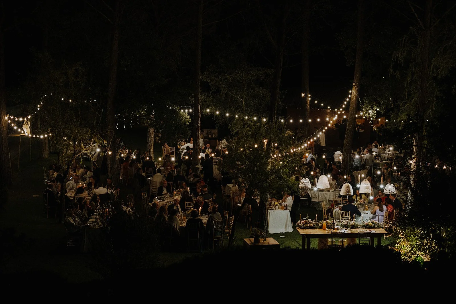 Nighttime outdoor wedding reception with strings of hanging lights, guests seated at tables in a wooded area.