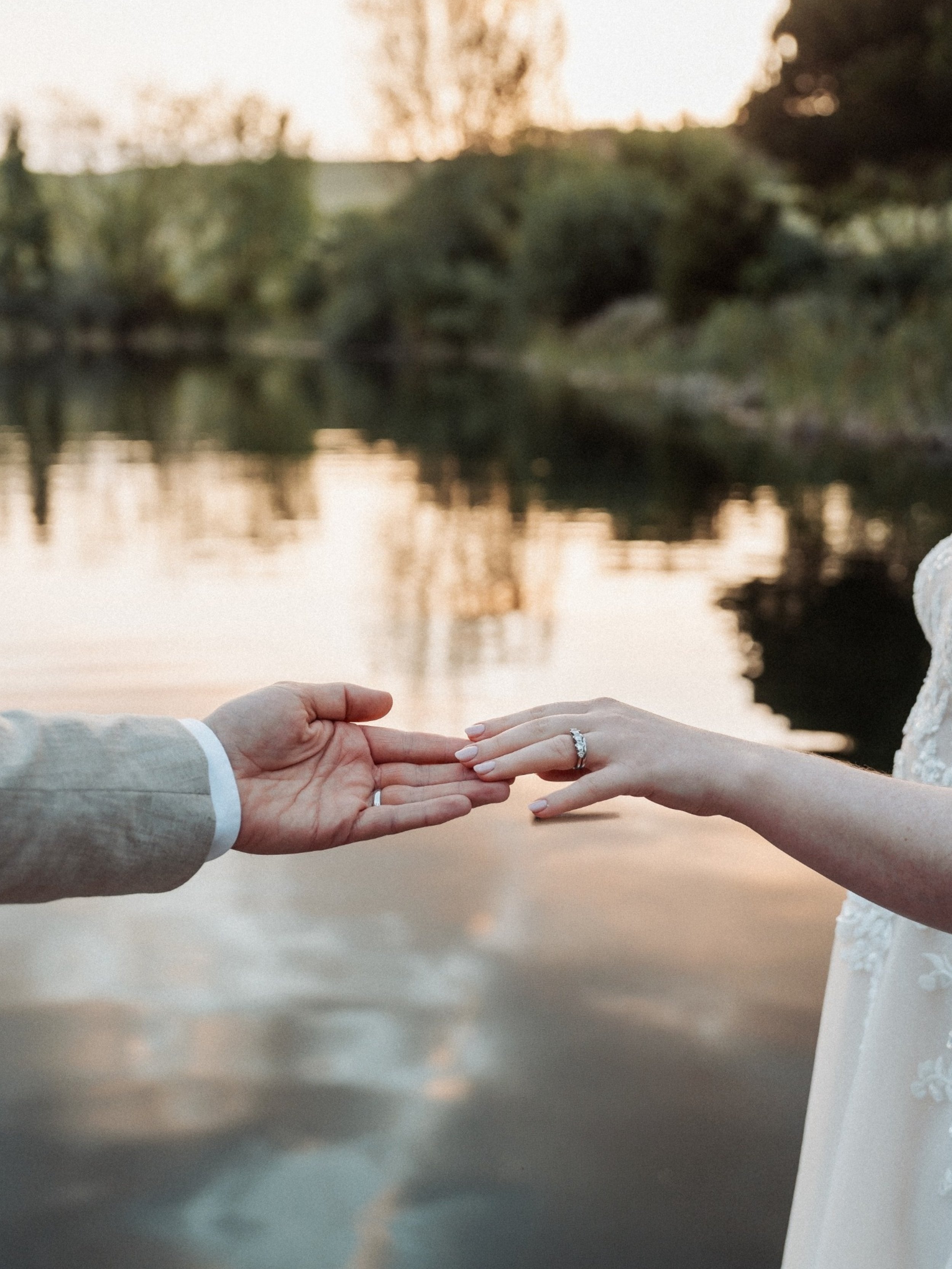 A bride and groom holding hands with a river and trees in the background during sunset, with the groom's hand near the bride's, showing her engagement ring.