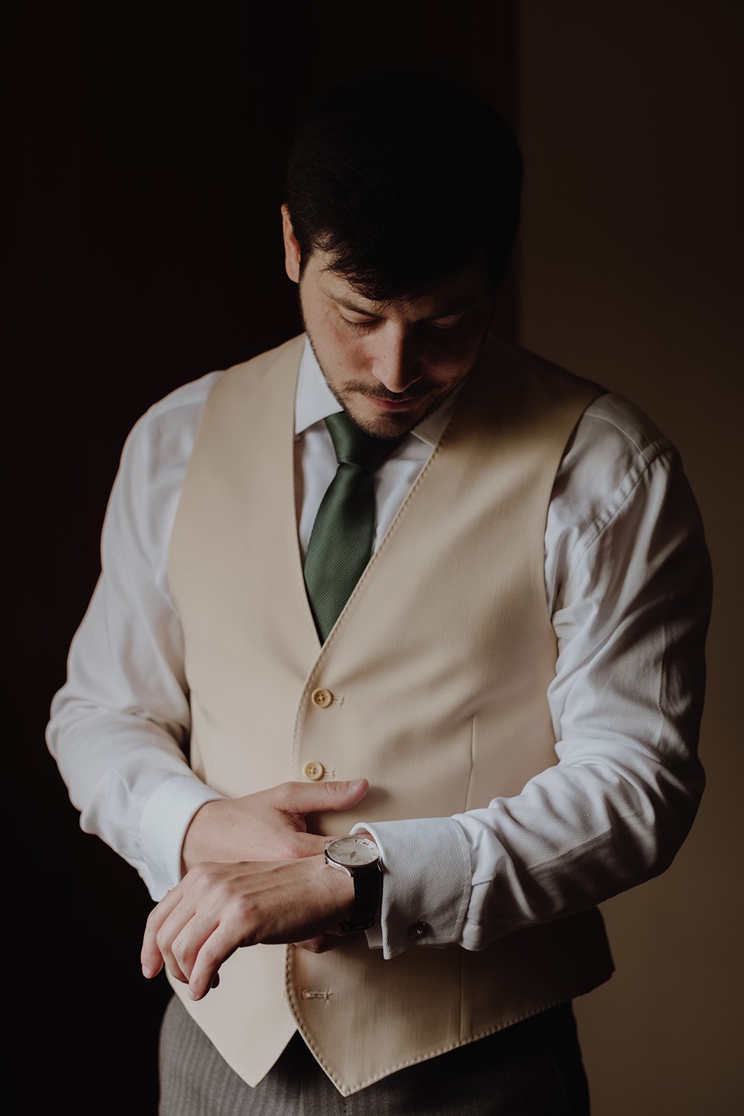 A man in a white dress shirt, beige vest, green tie, and watch adjusting his cuff in a dimly lit room.