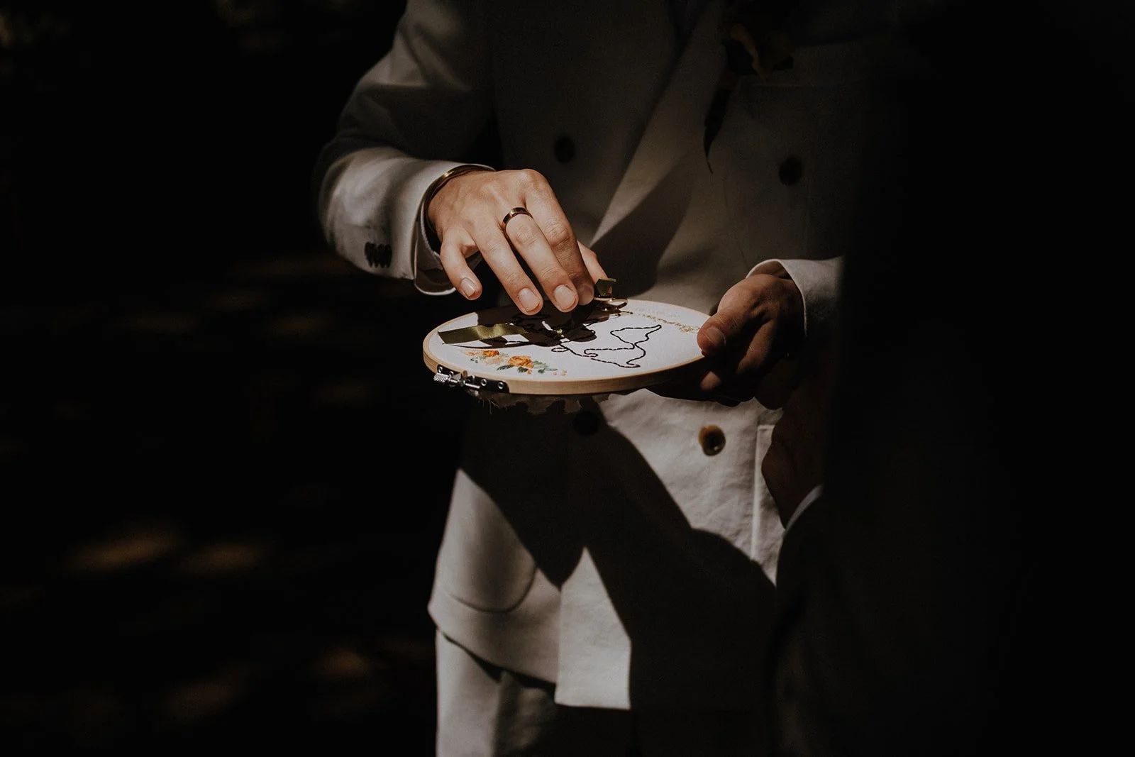 A person in a white suit holding and working on an embroidery hoop with a dark background.