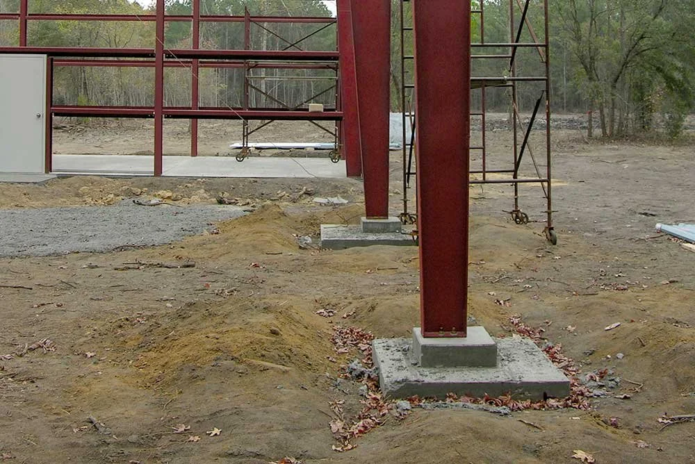 Construction site with red steel beams and scaffolding, dirt ground with small piles of dirt and scattered leaves.
