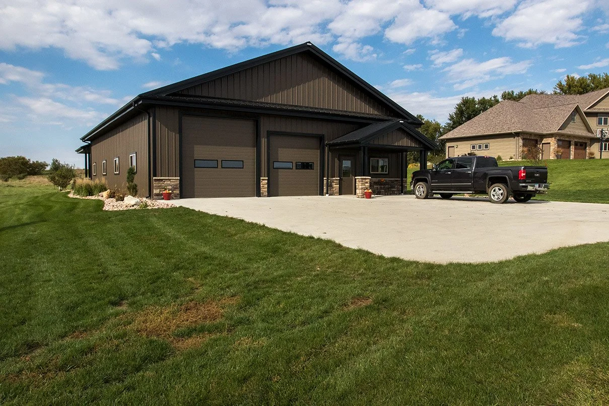 A large black metal garage with two doors, a small side porch, and a black pickup truck parked in front. Green grass surrounds a concrete driveway and neighboring homes are visible in the background under a partly cloudy sky.