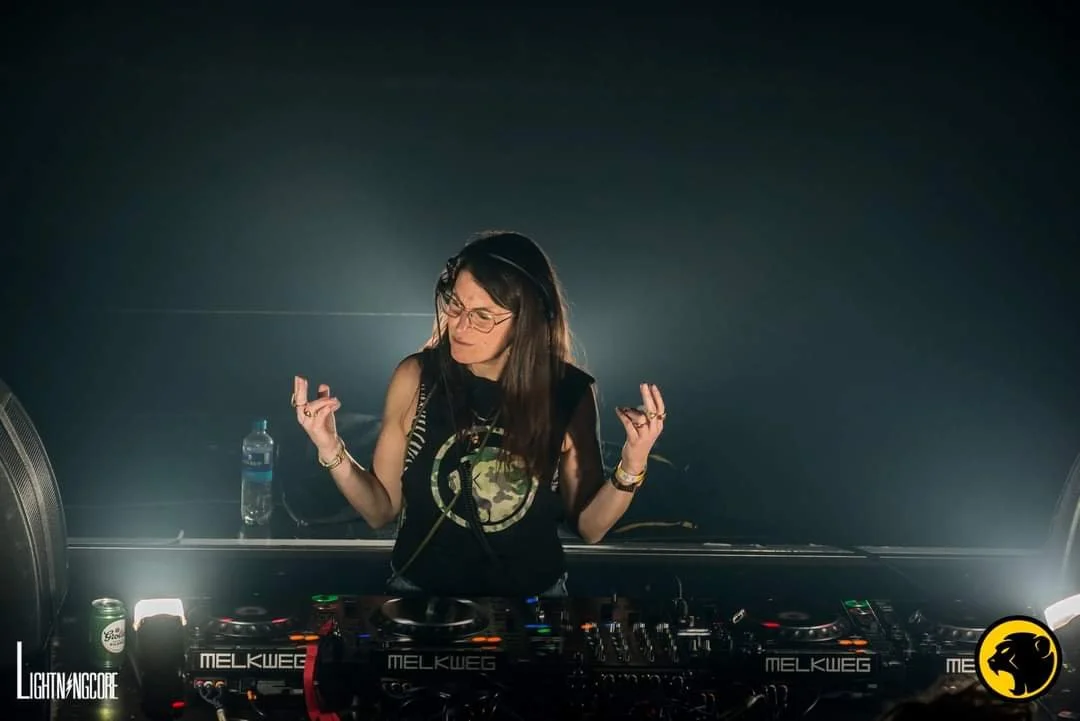 A woman DJing at a nightclub with DJ equipment, glasses, a water bottle, and a can of drink in front of her, with a dark background and spotlight. Adi-J wears her custom camo Cheeky Monday t-shirt