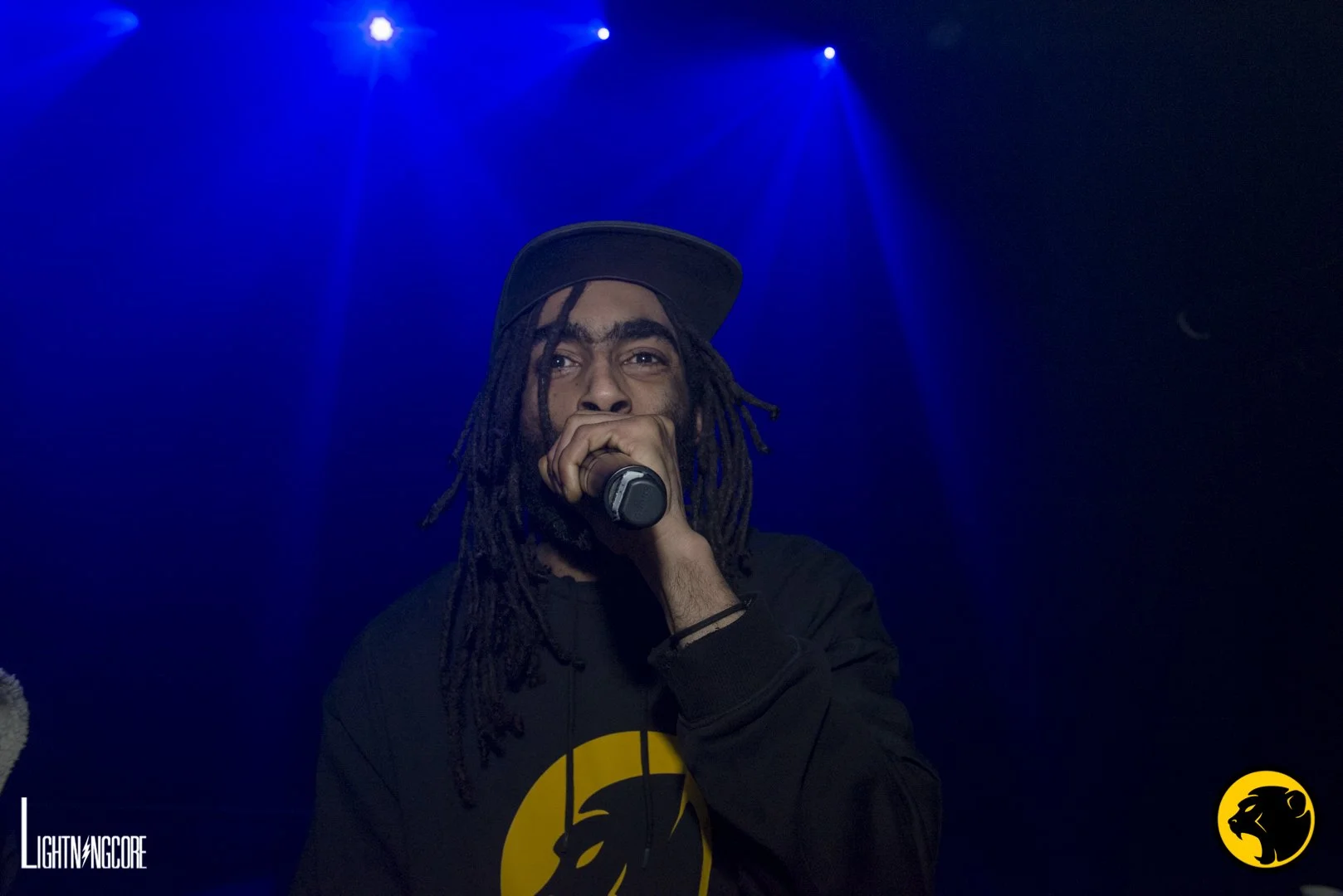 A man with dreadlocks wearing a black cap and black sweatshirt with a yellow Cheeky Monday logo, holding a microphone, on a stage with blue lighting.