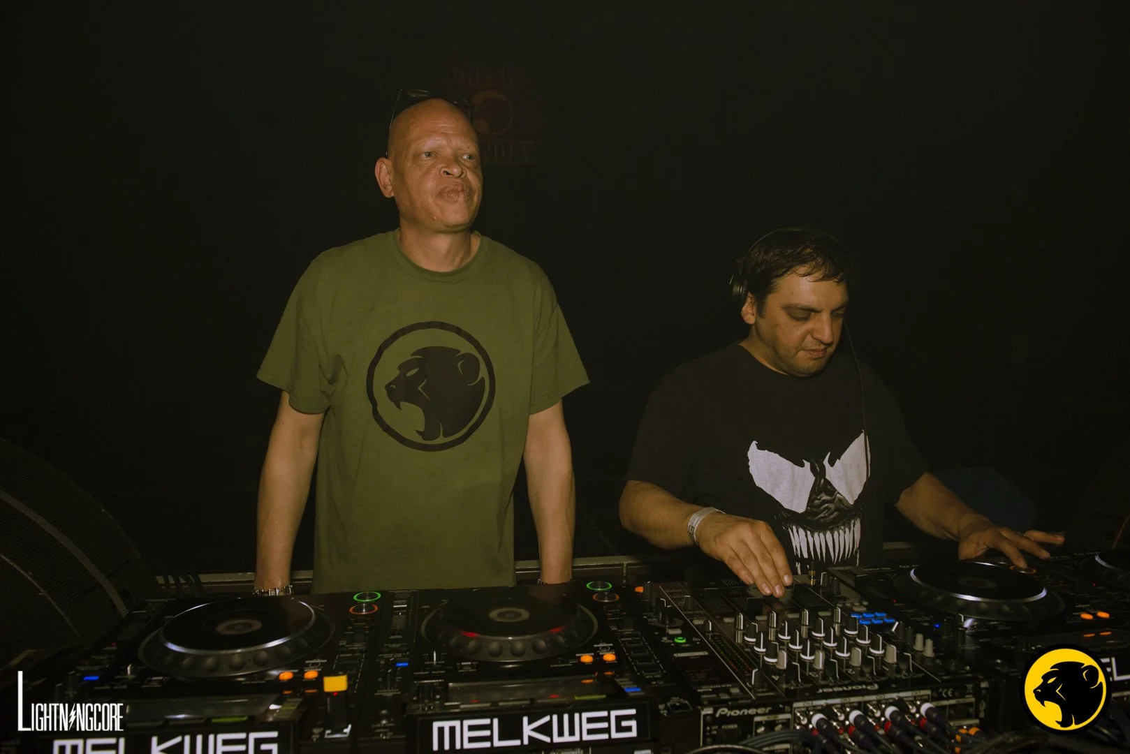 Bass80 and Insom. Two male DJs performing at a music event with a DJ mixer and turntables, black background. Bass80 wearing green Cheeky Monday t-shirt.