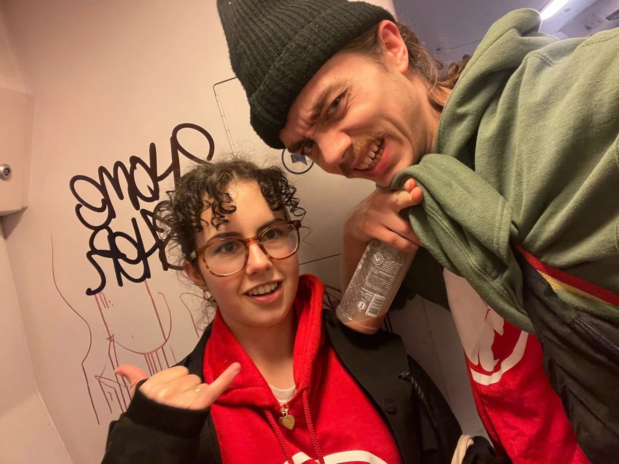 Two young adults taking a selfie in front of a wall with black graffiti and a drawing of a face mask. The woman and the man wear Cheeky Monday merch.