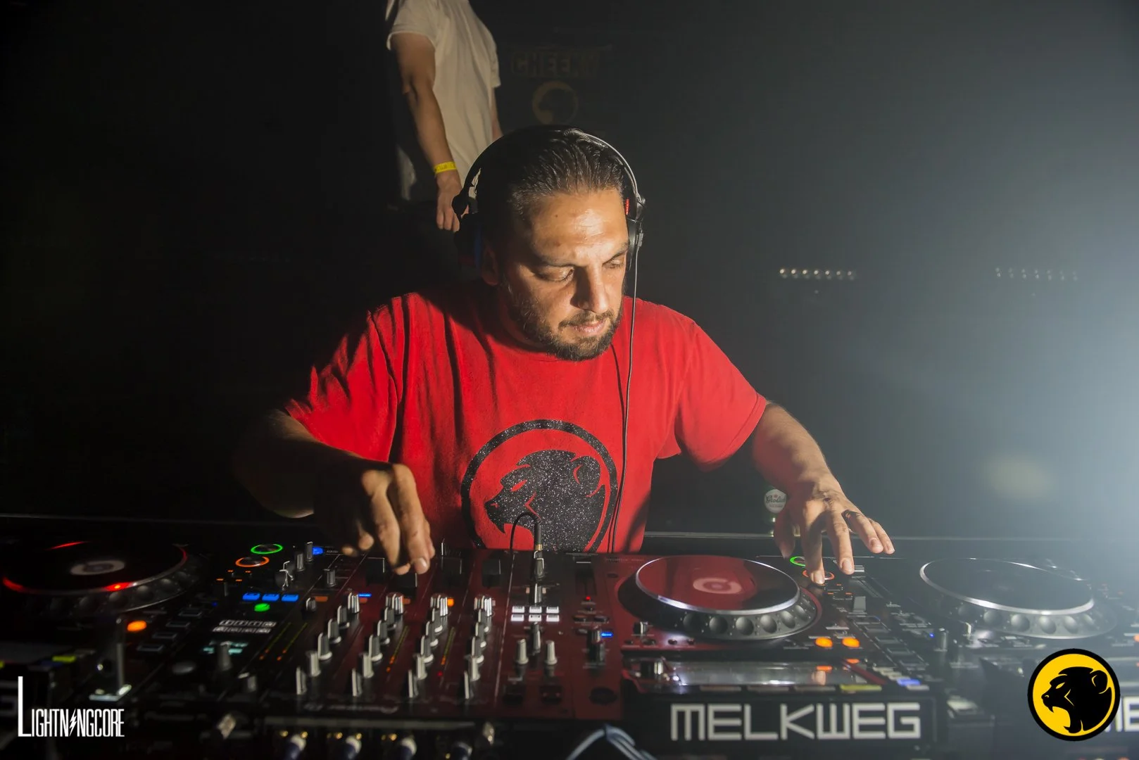 A DJ with long dark hair and a beard, wearing a red t-shirt with a Cheeky Monday logo, DJing at a club or event with a dark background and some light effects.