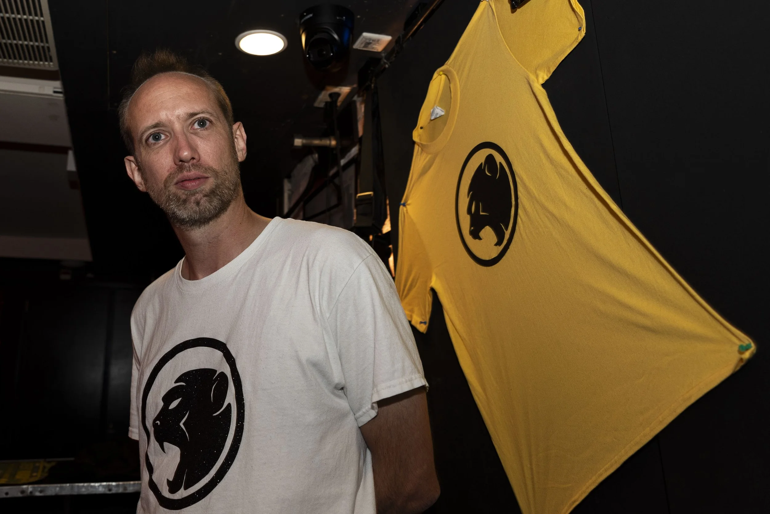 A man with light skin, light brown hair, and a beard wears a white t-shirt with a black Cheeky Monday logo. Behind him, a yellow flag with the same lion logo is displayed on a black background.
