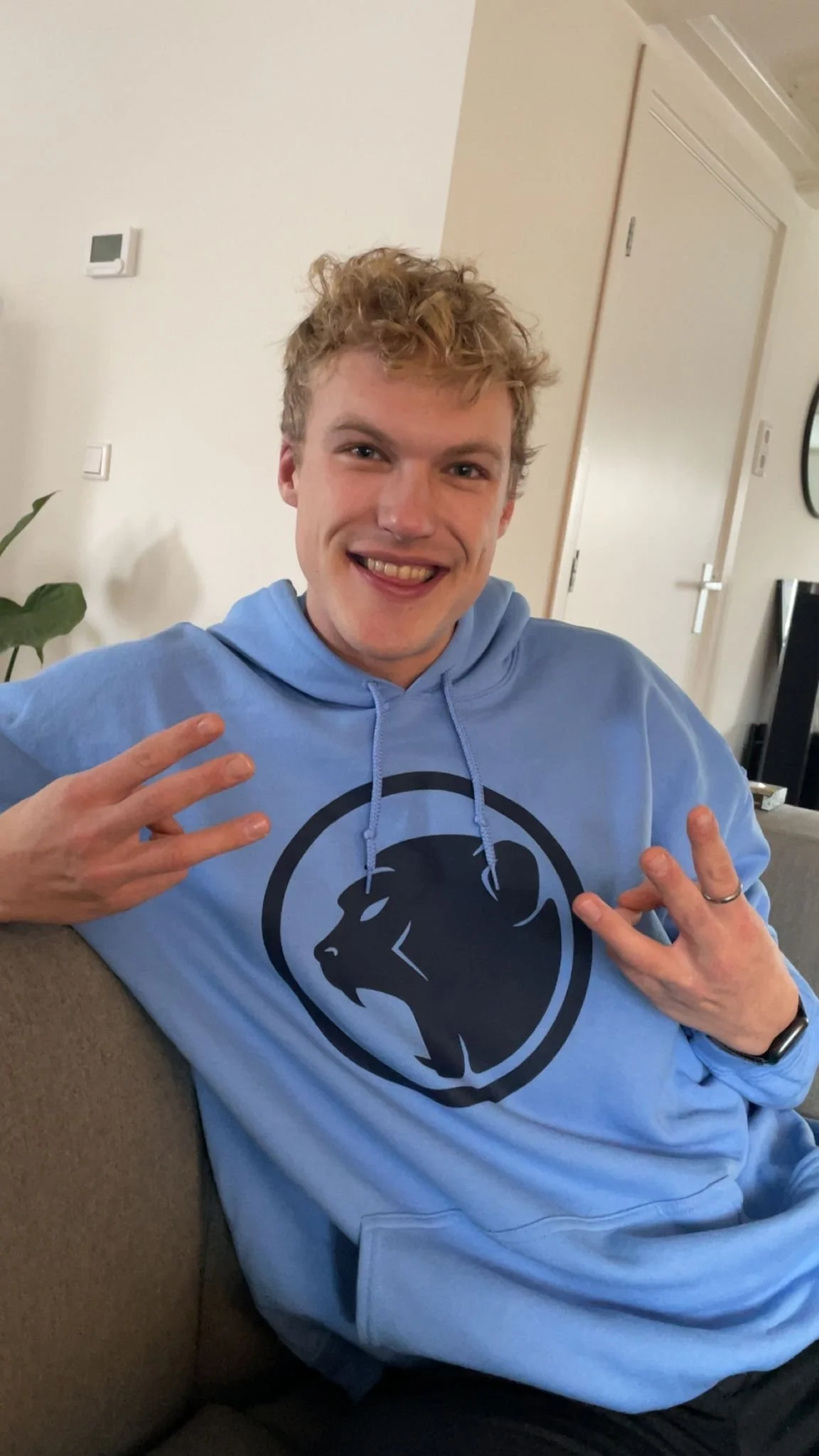 A young man with curly blond hair smiling and making a peace sign and a hand gesture while sitting on a couch, wearing a light blue hoodie with a black Cheeky Monday logo on it in a living room.