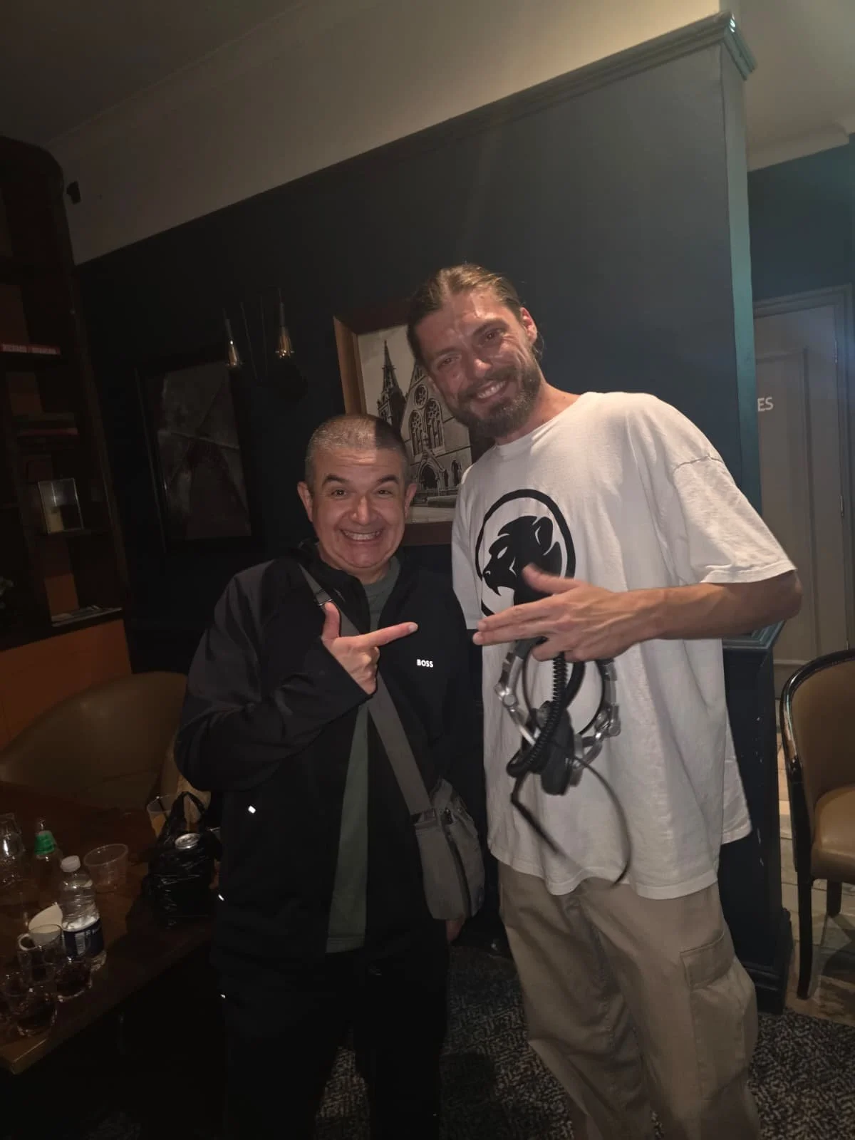Two men smiling and posing for a photo indoors, one wearing a black jacket and the other wearing a white t-shirt with a black Cheeky Monday monkey logo, holding headphones.