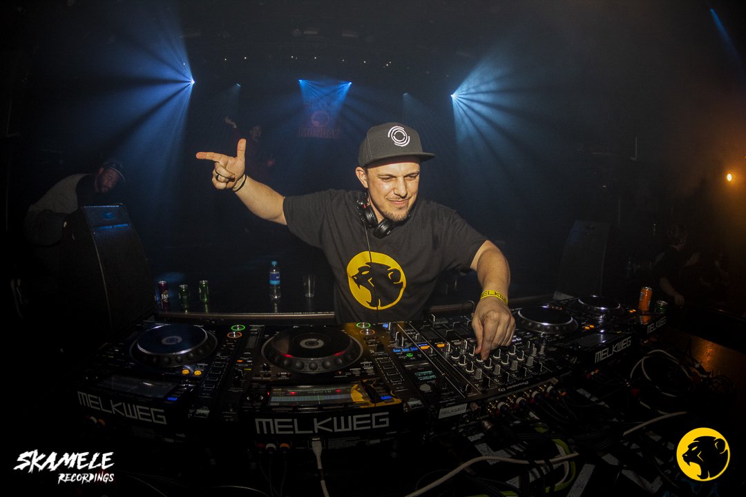 A DJ wearing a black black cap and black and yellow Cheeky Monday t-shirt, with headphones around his neck, is DJing at a nightclub, with blue and orange lighting and a crowded dance floor.