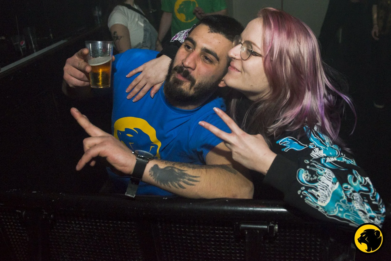 A man with a beard and a woman with dyed pink hair are posing together at a social event. The man is holding a drink, and the woman is making a peace sign with her hand. They look happy and are close to each other. Blue Cheeky Monday shirt.
