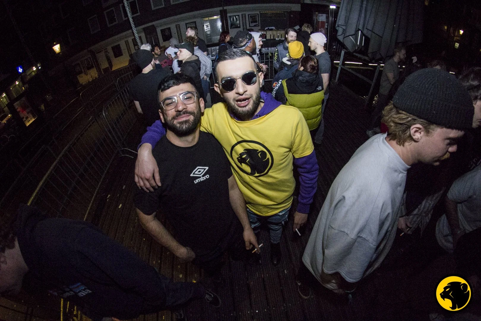 A group of young people at a Cheeky Monday, with two men in the center posing and wearing glasses, one in a black t-shirt and the other in a yellow Cheeky Monday t-shirt , on a rooftop with several others in the background.