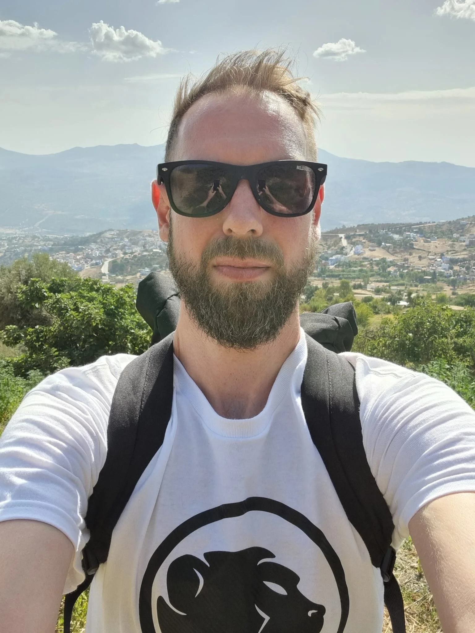 A man with a beard and sunglasses and Cheeky Monday t-shirt taking a selfie outdoors, with a backpack on his shoulders. The background features a landscape of trees, hills, and houses under a partly cloudy sky.