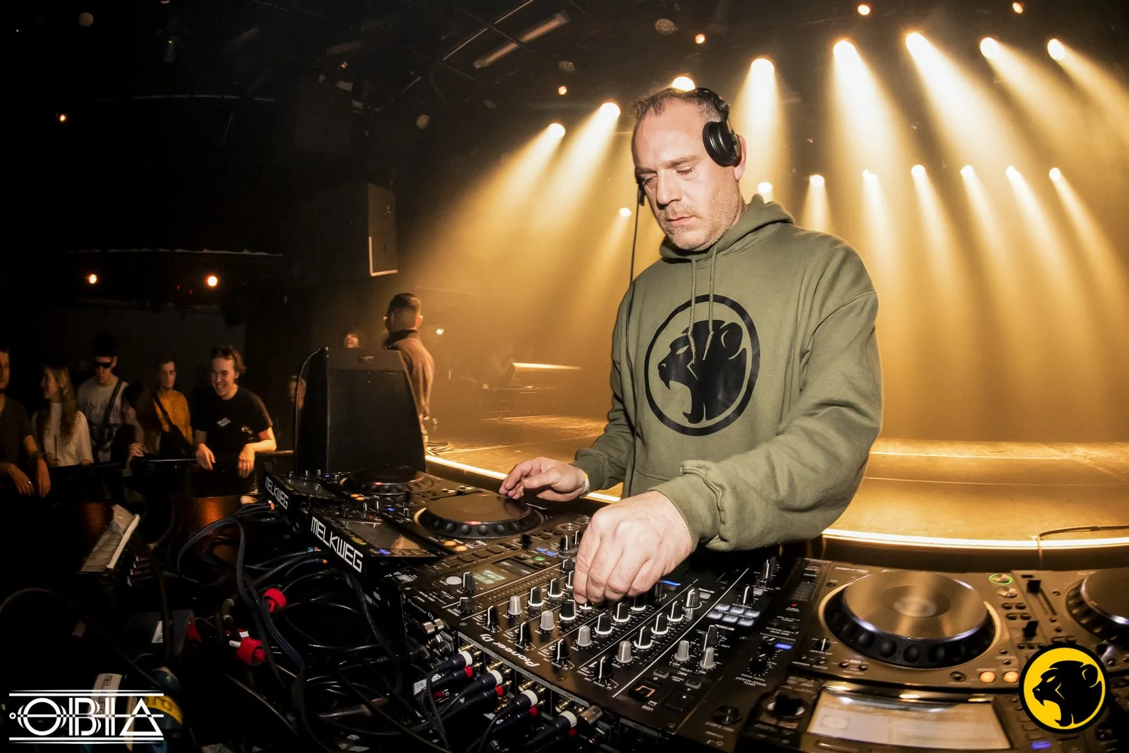 Loco, A DJ wearing a green Cheeky Monday hoodie with a monkey logo, using DJ equipment on stage at a nightclub with yellow stage lights and an audience in the background.