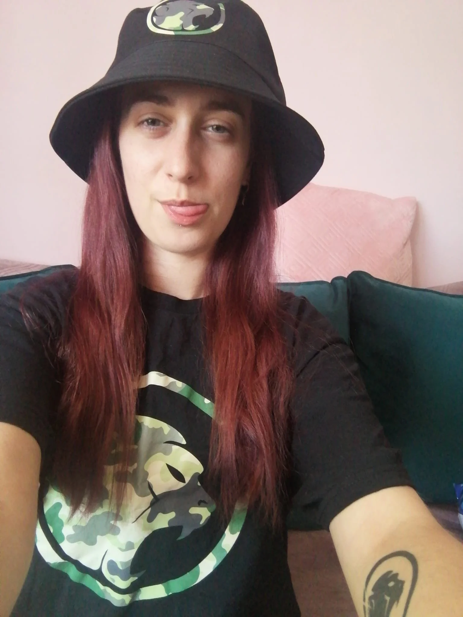 A person with long reddish-brown hair, wearing a black bucket hat with a Cheeky Monday logo, a black T-shirt with a logo, and showing a tattoo on her forearm. She is sticking out her tongue slightly.