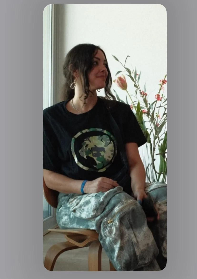 A woman with dark wavy hair sitting indoors near a window with a flower arrangement in the background. She is wearing a black Cheeky Monday t-shirt with a camouflage design and camouflage pants, and is holding a phone in her lap.