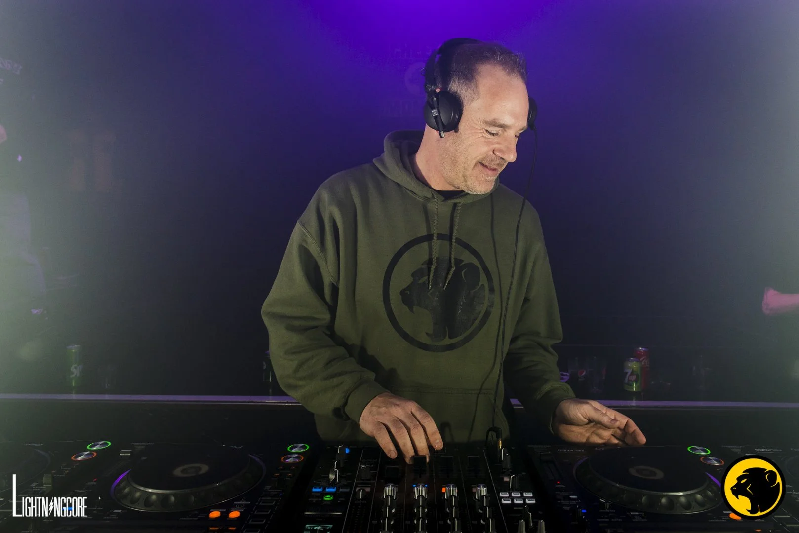 Loco A DJ wearing a green Cheeky Monday hoodie with a monkey logo, listening to music through headphones, standing behind a DJ setup with turntables and mixer in a dark venue.