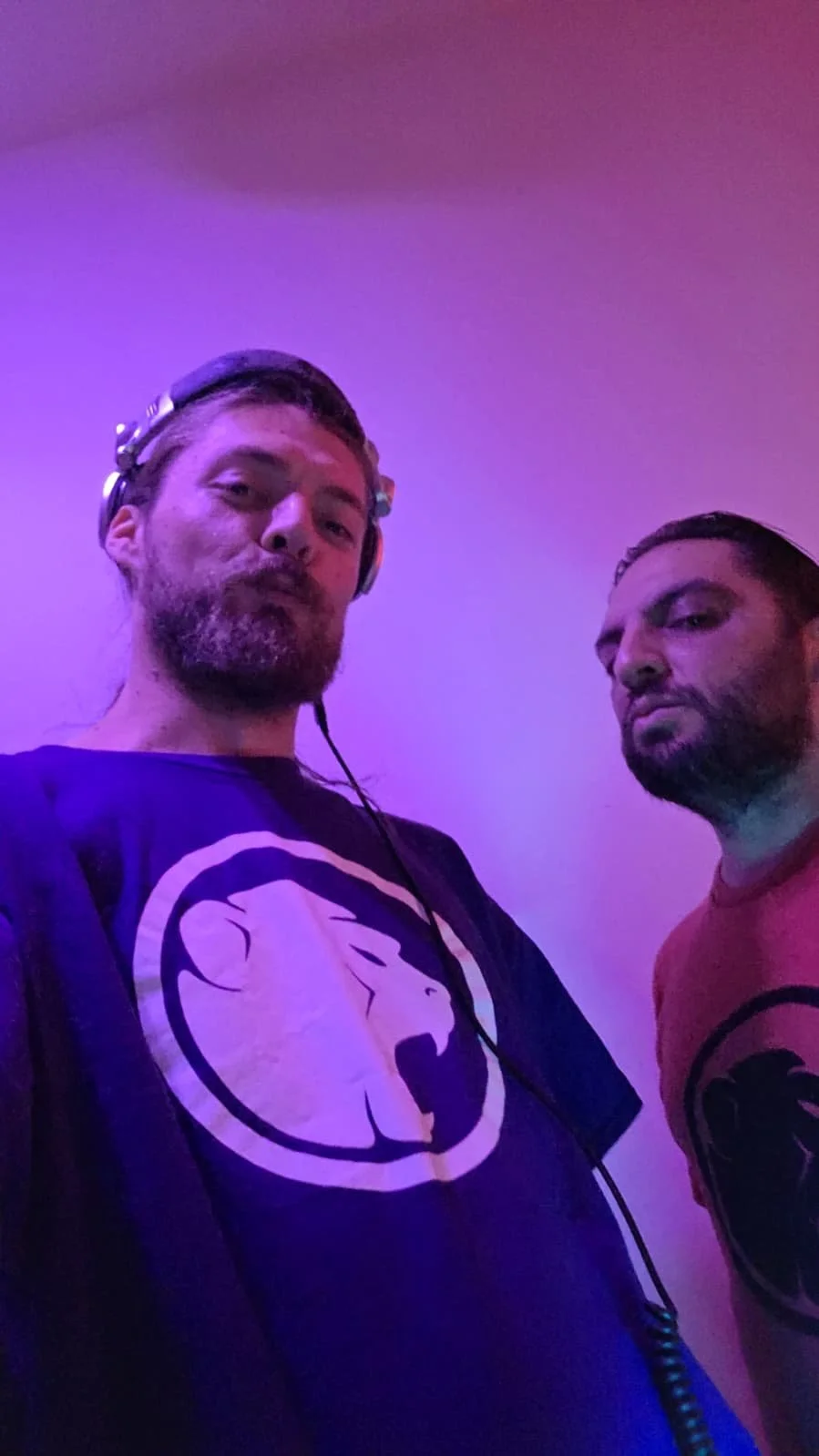 Two men are posing together with purple and pink lighting, both wearing shirts with Cheeky Monday logos, one with headphones around his neck.