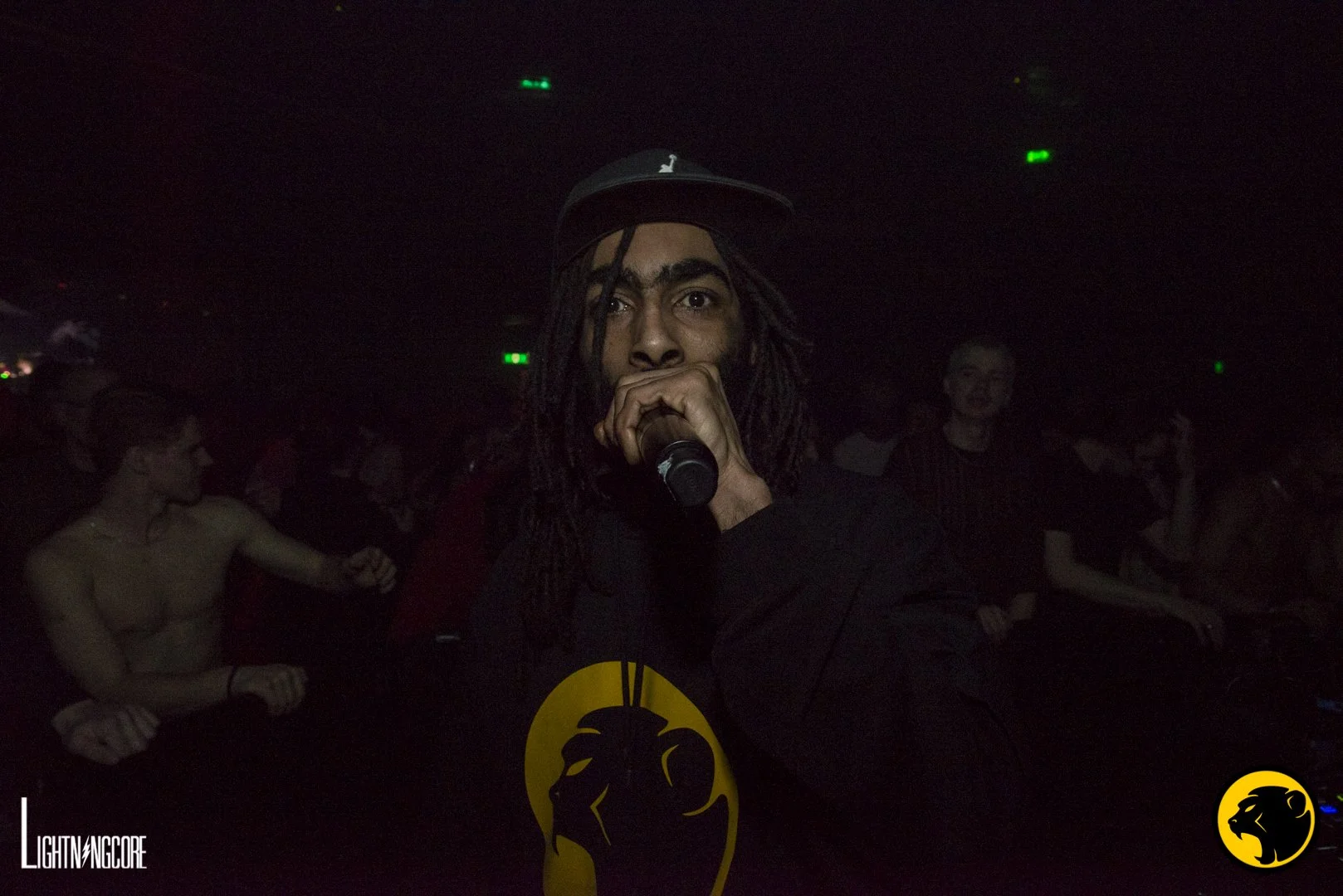 A DJ with dreadlocks and a black cap holding a microphone in a dark nightclub with people dancing in the background. Cheeky Monday hoodie.