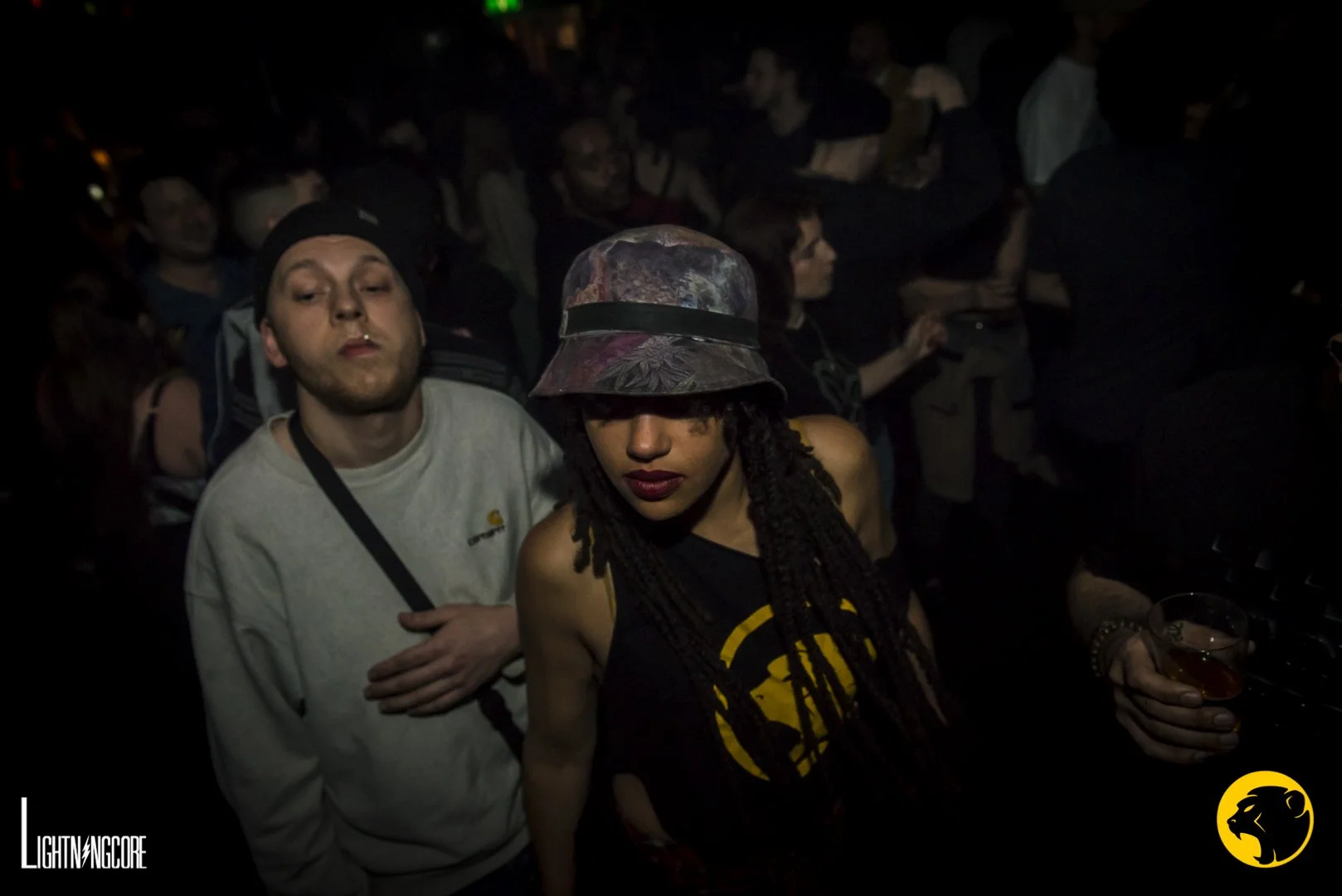 Crowd of people at a dark concert or nightclub, some with drinks, some with headphones, dark lighting, two individuals in the foreground, one with a bucket hat, Cheeky Monday vest and dreadlocks, and another with a beanie and a nasal cannula.