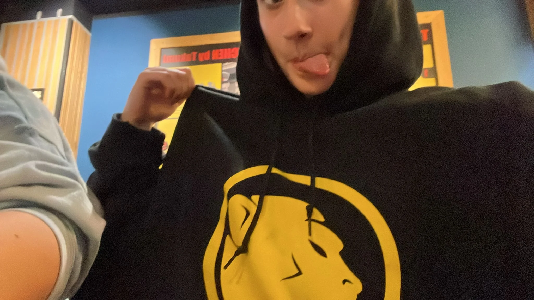 Person wearing a black hoodie with a yellow Cheeky Monday logo making a playful face with tongue out in an indoor setting with bright-colored walls and posters.