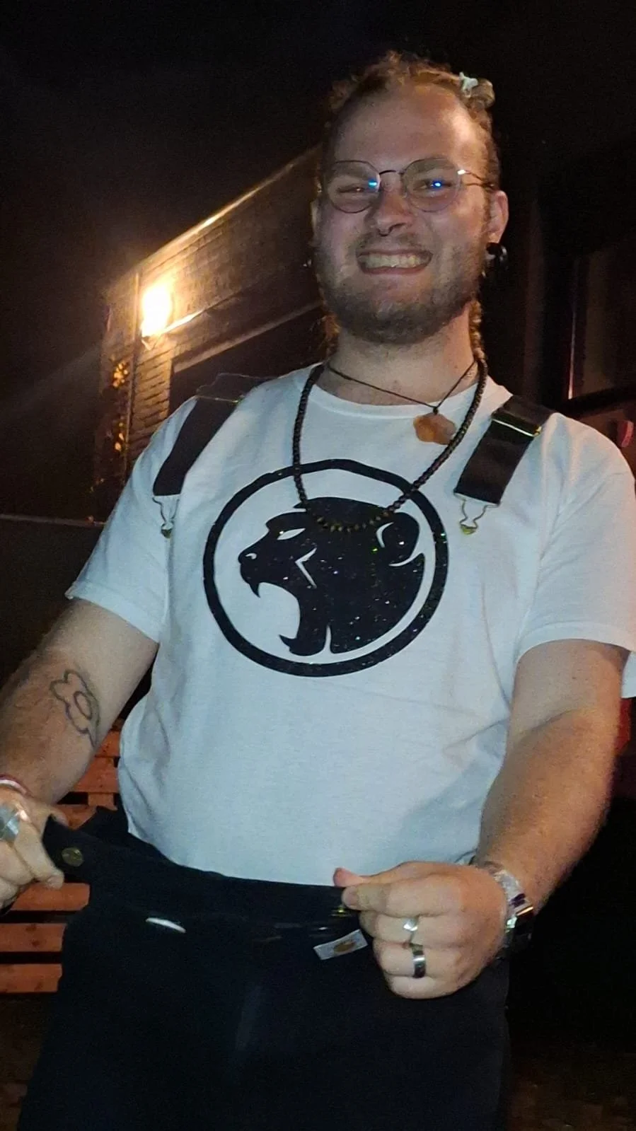 A smiling young man with glasses, tattoos, and a beard, wearing a white T-shirt with a black Cheeky Monday logo, a necklace, and rings, standing outdoors at night near a lit brick building.