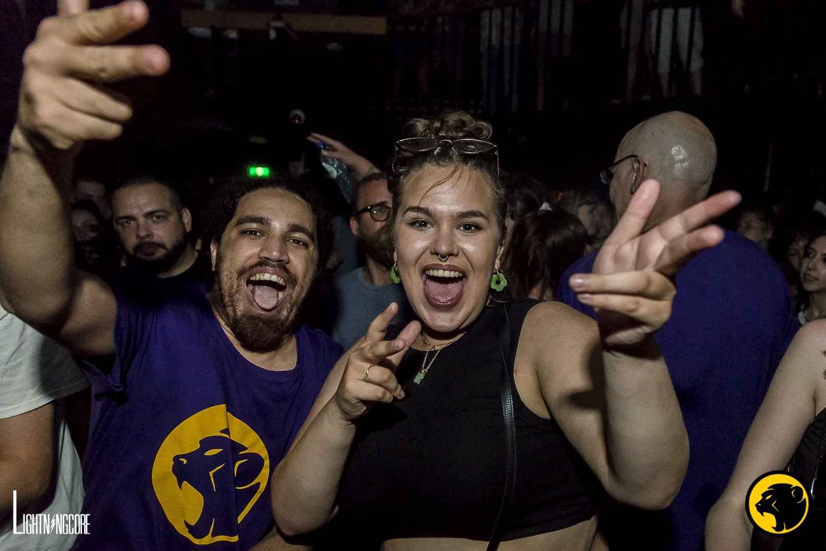 Group of people at a concert or party, smiling and making peace signs, with a woman in the center and a man on her left, all facing the camera. Purple Cheeky Monday shirt.