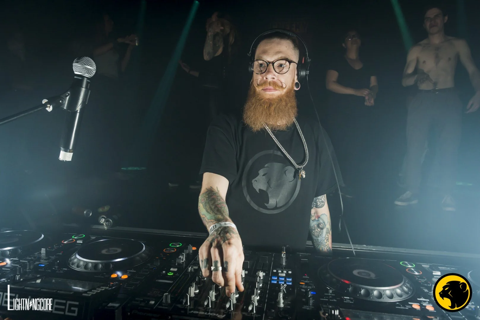 Multiplex. A bearded DJ wearing glasses, headphones, and a black T-shirt with a Cheeky Monday monkey logo, mixing music at a nightclub with people dancing in the background.