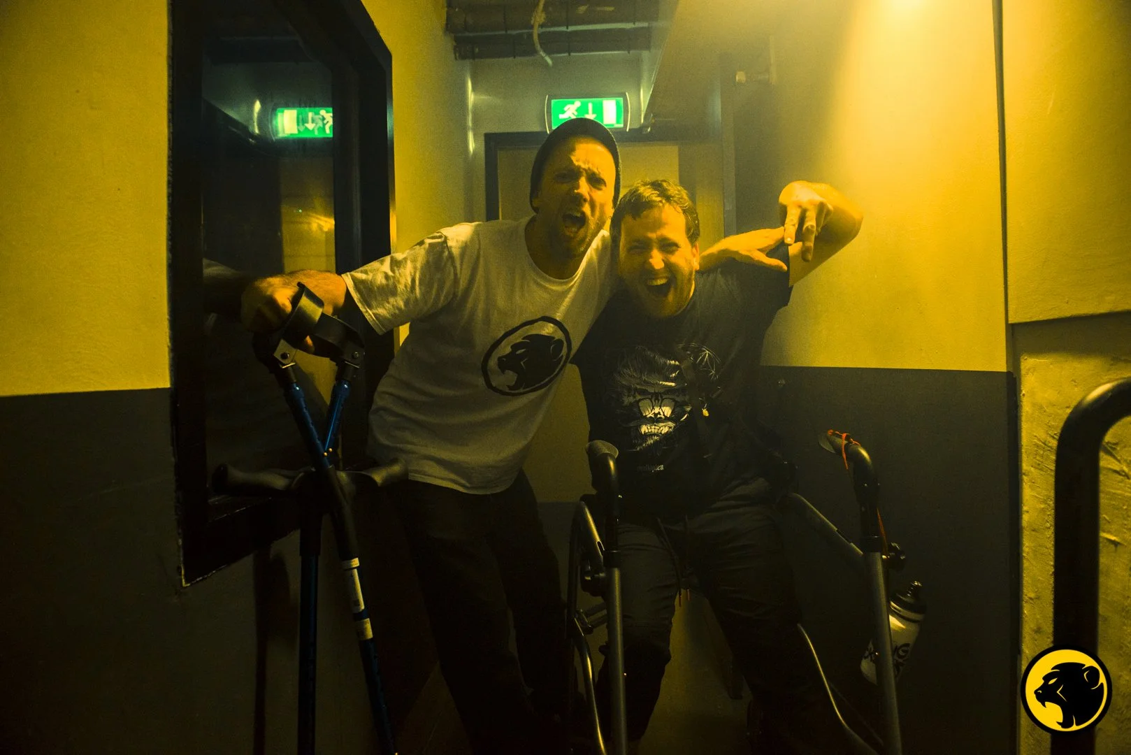 Two men with disabilities and joyful expressions posing together in a dimly lit hallway, each holding a walker, with emergency exit signs above. Matching Cheeky Monday merch.