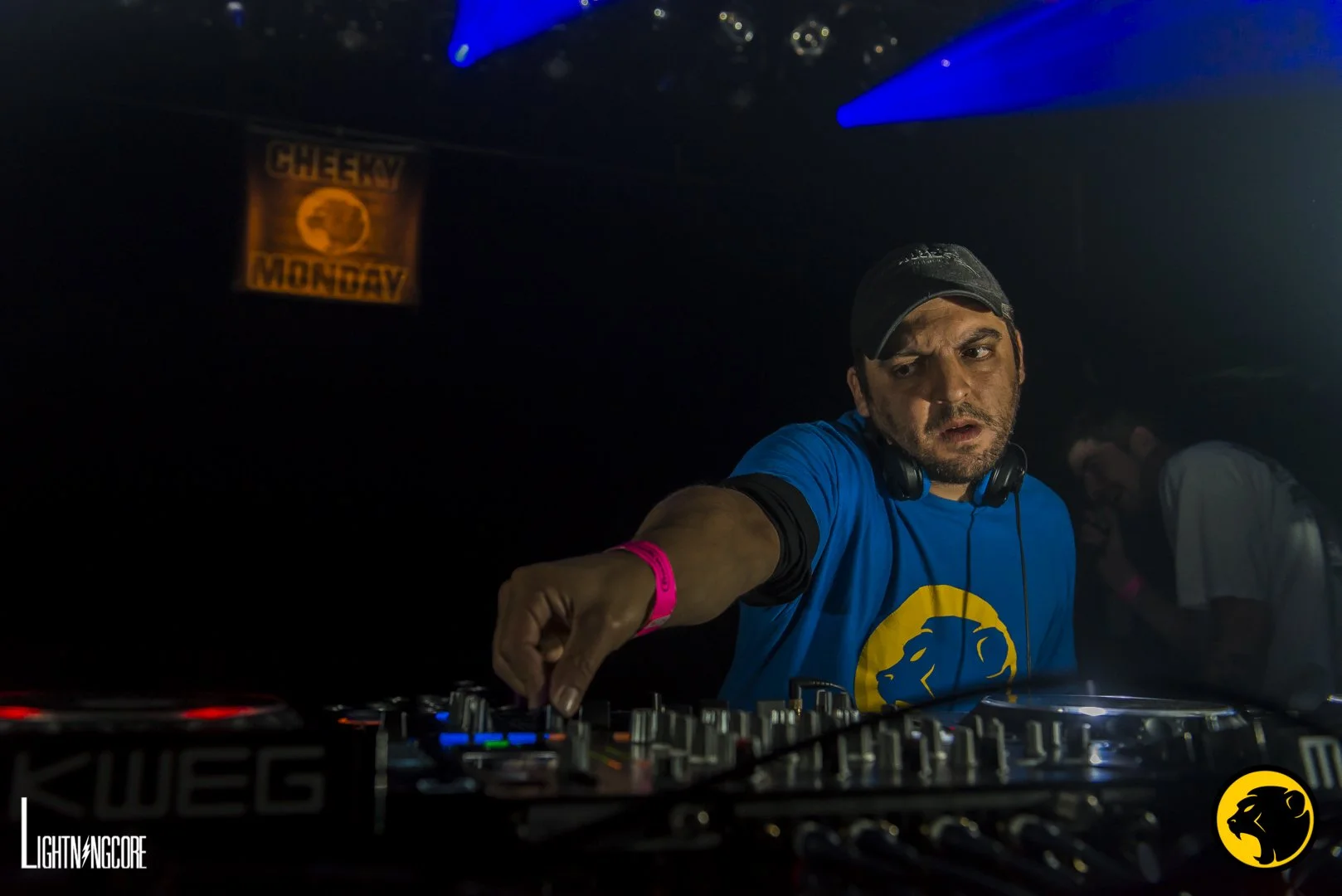DJ wearing a blue Cheeky Monday logo shirt and black cap, adjusting audio equipment in a dark club with blue and purple lights.