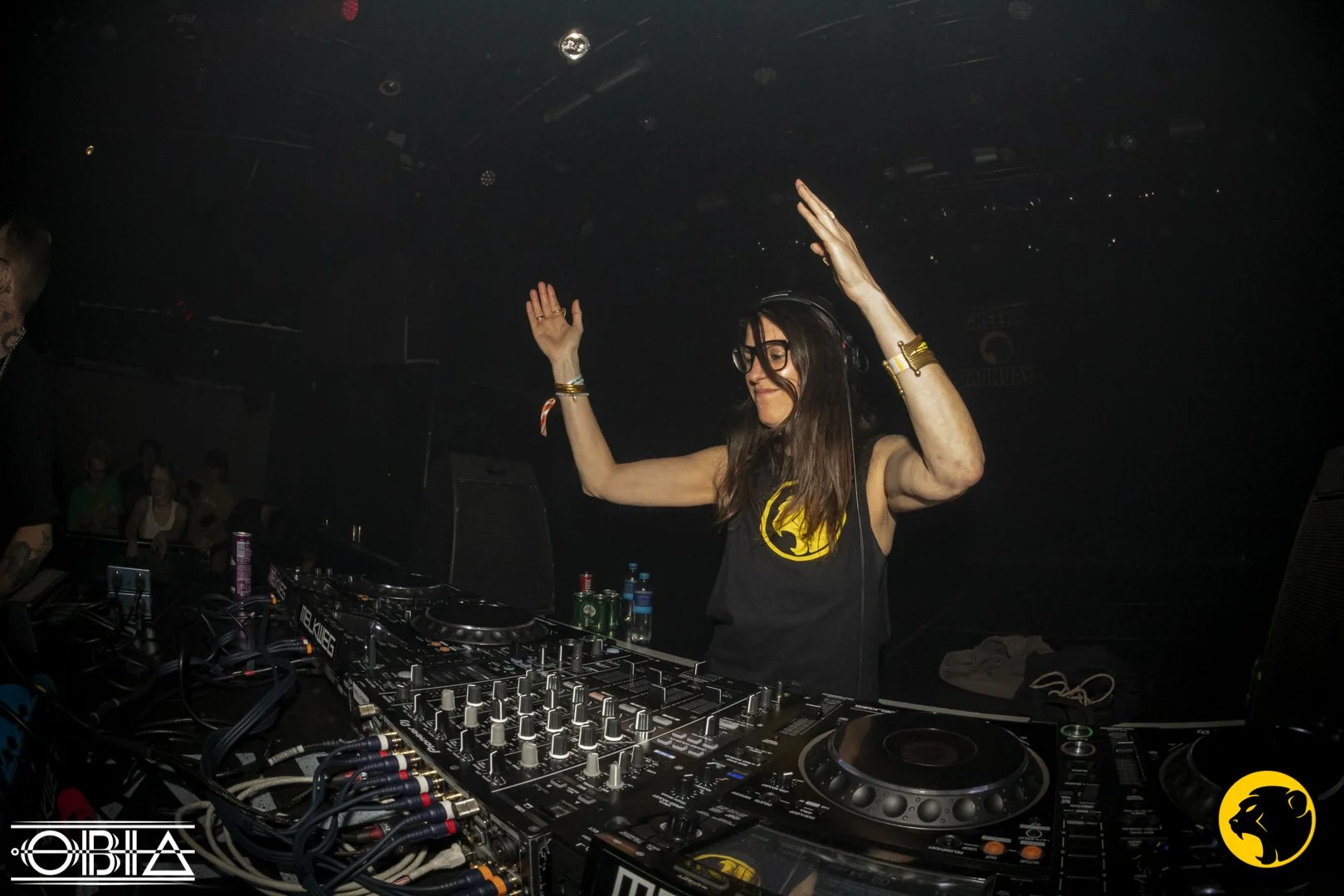 Female DJ wearing glasses and a black t-shirt with a yellow Cheeky Monday logo, standing behind DJ equipment with arms raised, performing at a club or music event.