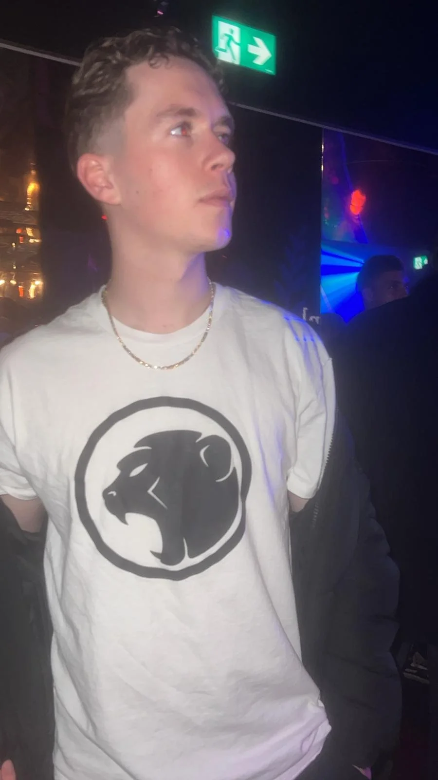 Young man wearing a white T-shirt with a Cheeky Monday graphic, gold chain necklace, in a dimly lit nightclub with a green exit sign overhead.