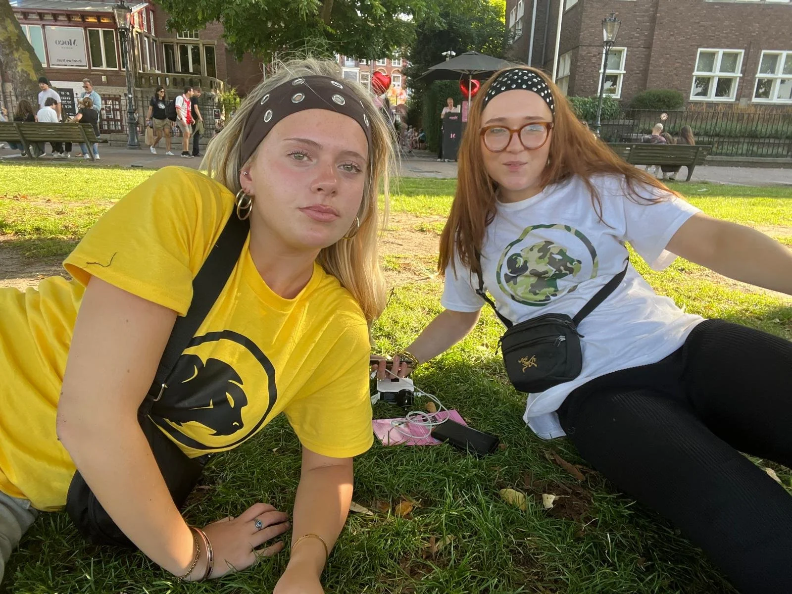 Two young women sitting on the grass in a park, wearing matching Cheeky Monday logo t-shirts. One is wearing a yellow shirt, the other white. They are surrounded by people, benches, trees, and buildings in the background.