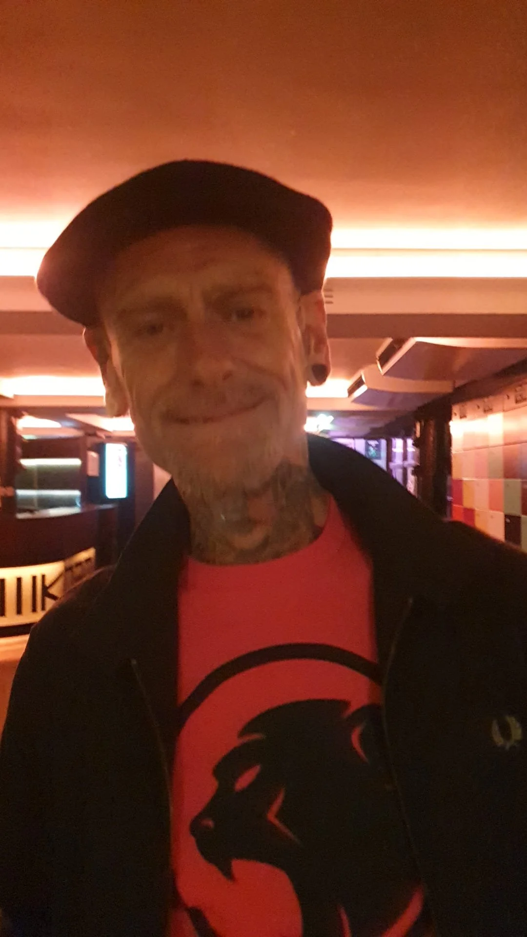 A man with tattoos on his face, neck, and ears, wearing a black cap, a black jacket, and a black and orange Cheeky Monday t-shirt, is smiling at the camera in a dimly lit indoor space.