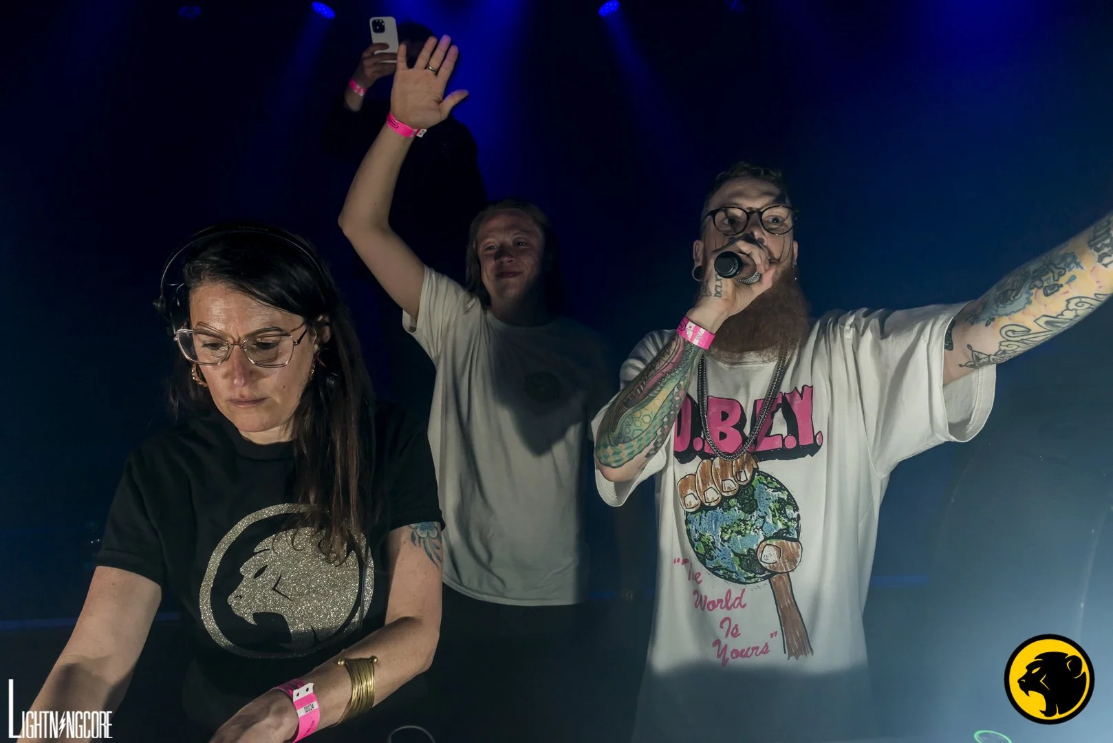 Three people at a DJ booth with one person singing into a microphone, a woman focused on mixing music, and a man raising his hand, all wearing pink wristbands, in a dark club or concert setting. Adi-J wears a silver sparkling Cheeky Monday logo.