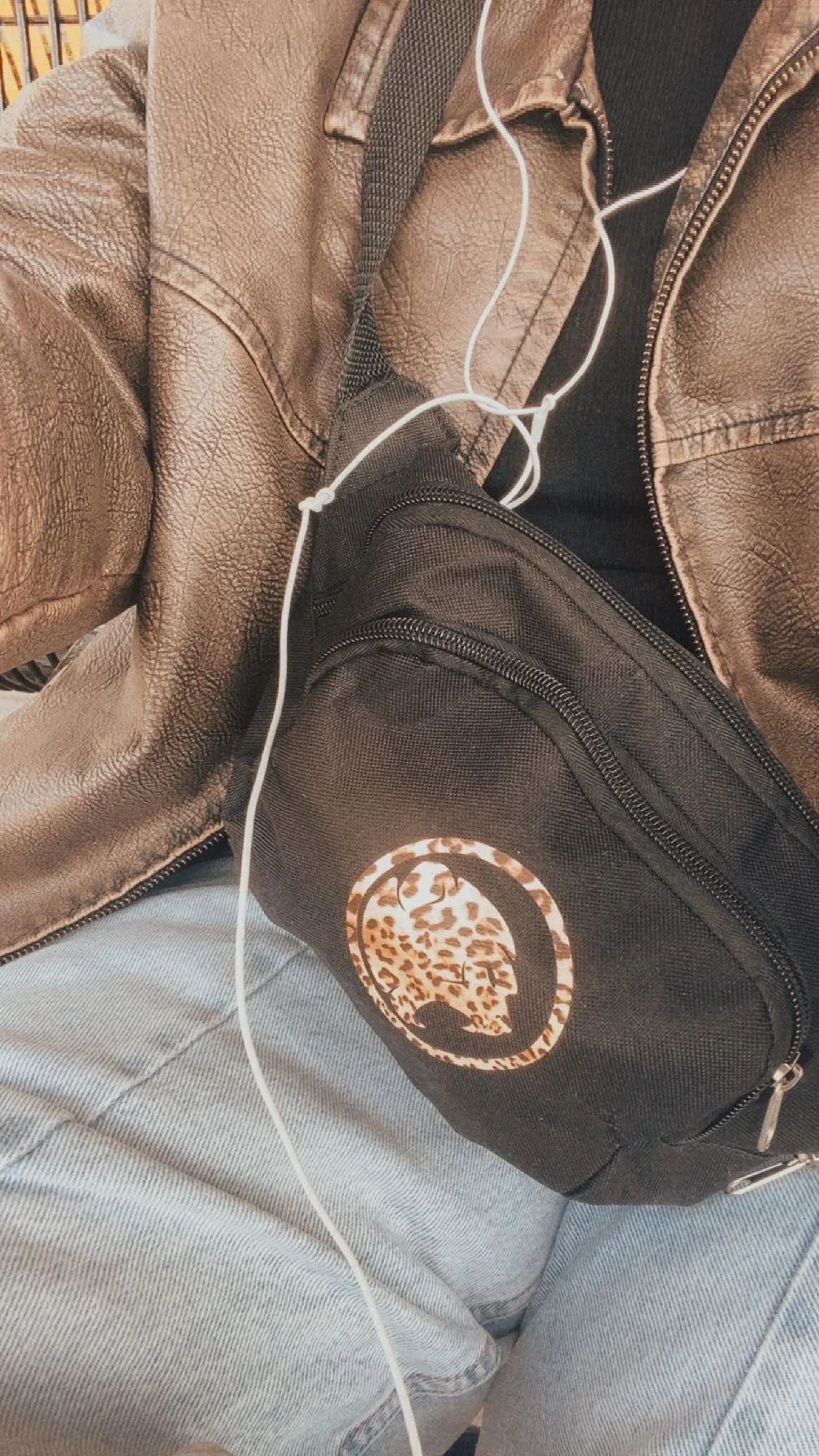 Close-up of a person wearing a brown leather jacket, black shirt, light blue jeans, black crossbody bag with a Cheeky Monday logo, and white earphones.