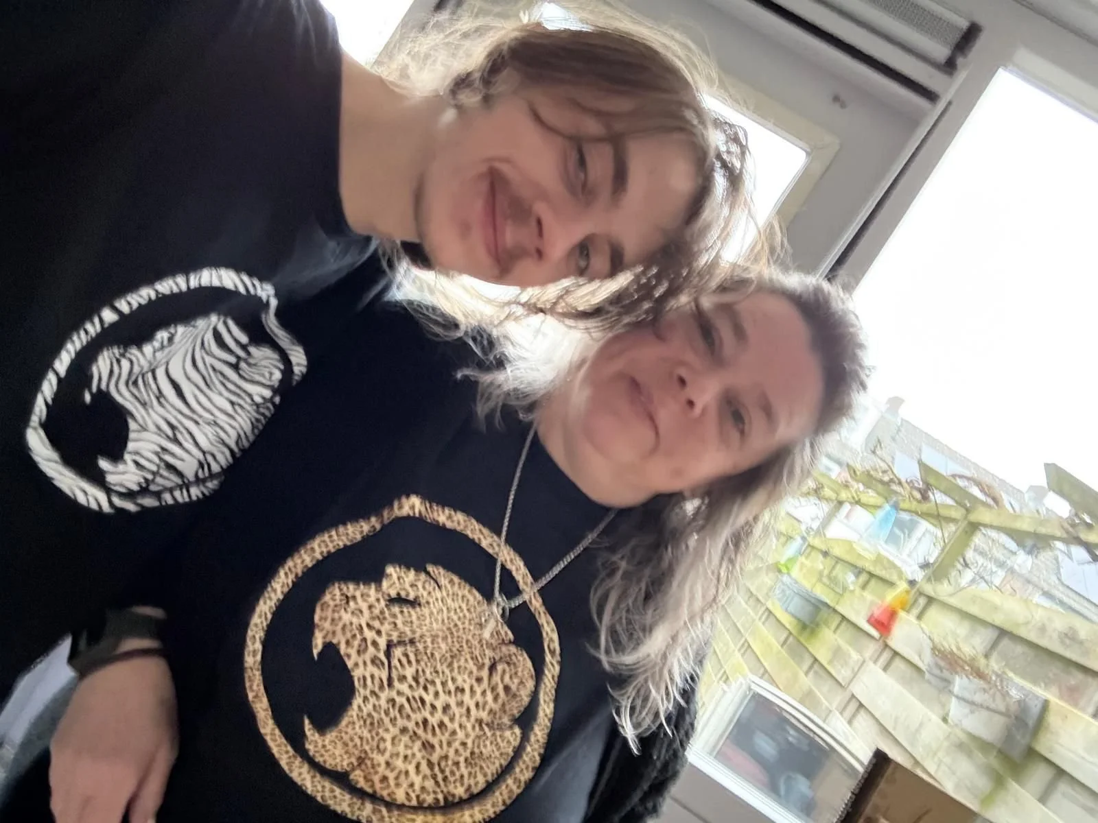 Two women with long, wavy hair smiling at the camera in a bright room with a window and outdoor view. Both wearing Cheeky Monday Tshirts.