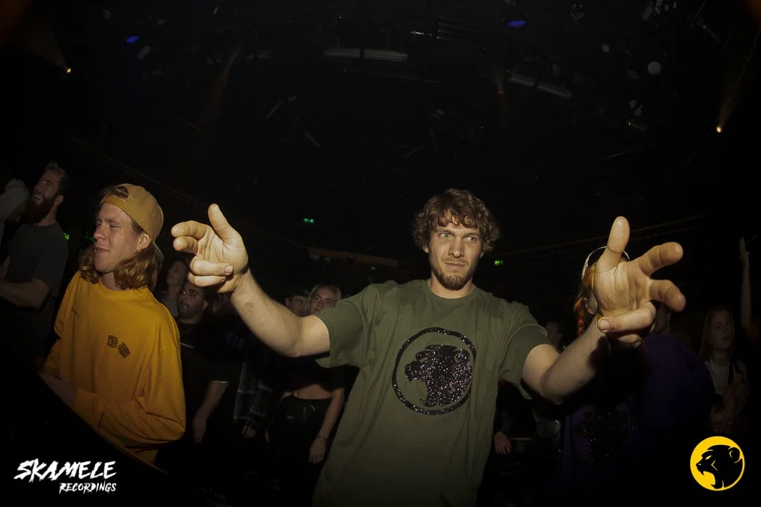 A man with curly hair and a beard wearing a green Cheeky Monday T-shirt making hand gestures at a concert or event. People are in the background, some with their hands raised, in a dark indoor setting with stage lighting.