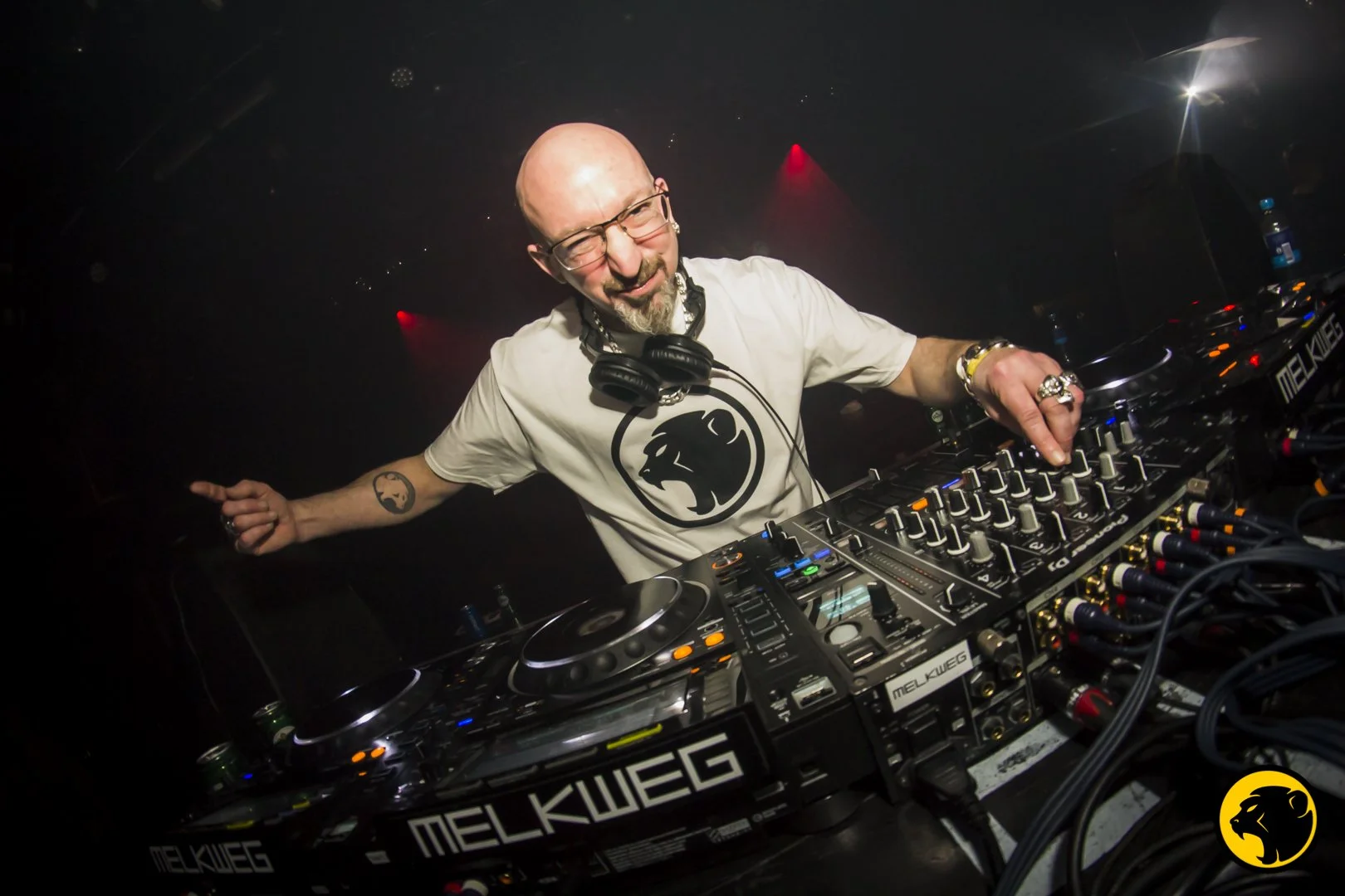 Sinistah A DJ with glasses, multiple rings, and tattoos on his arm, wearing a white t-shirt with a monkey logo, is behind a DJ console, smiling and pointing with one hand in a dark club with red and white lights.