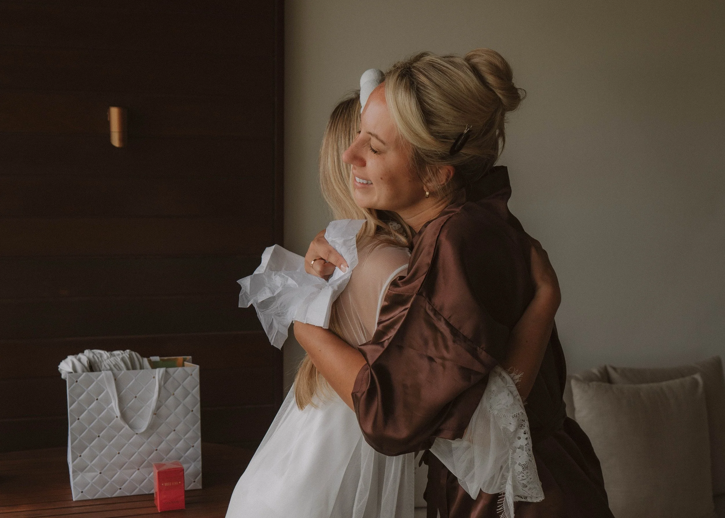 A woman hugging a young girl, both smiling, in a warmly lit room with a gift bag and small box on a wooden table nearby. yarra valley and Melbourne wedding photographer
