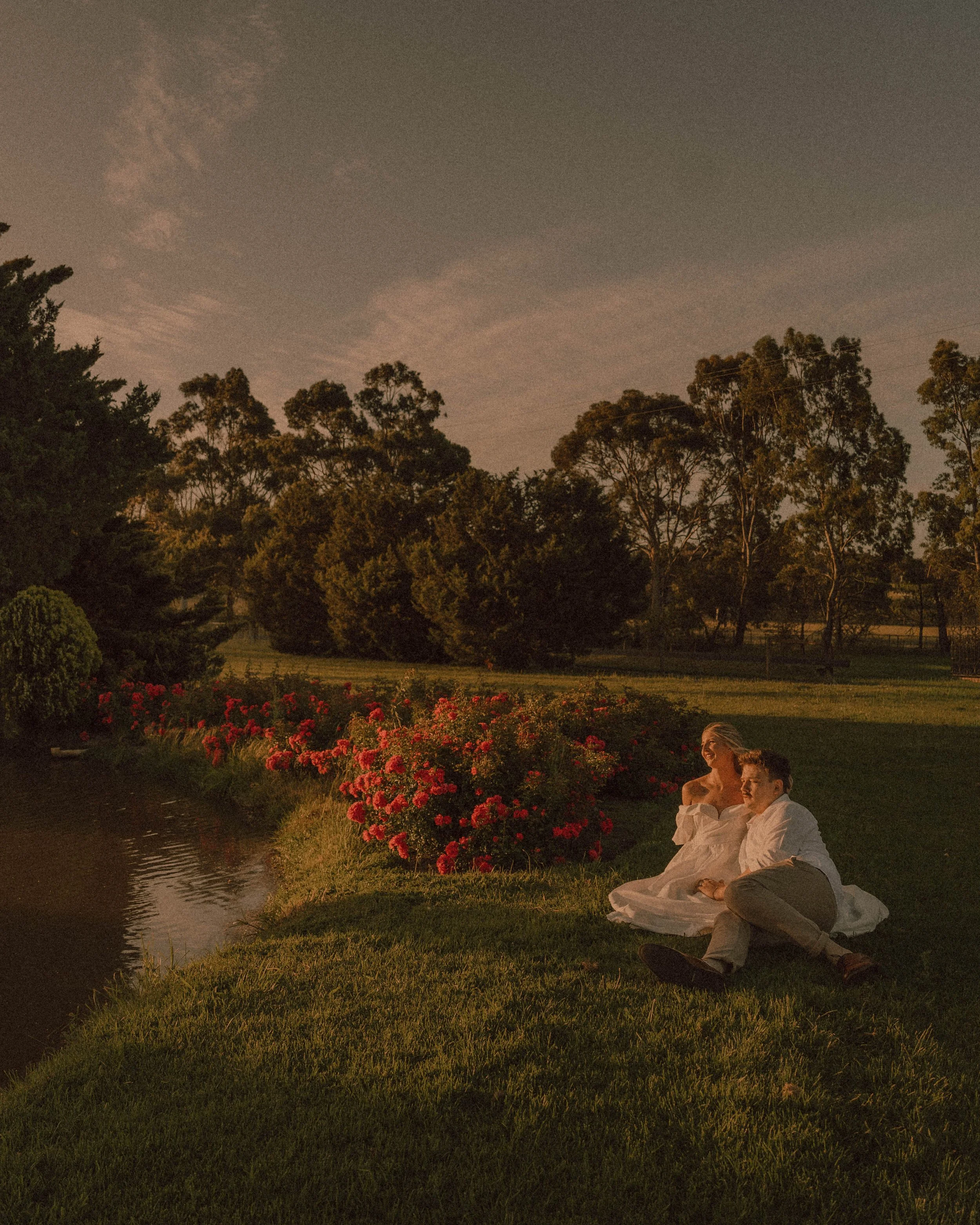 A couple sitting on the grass by a pond during sunset, with trees and bushes in the background and colorful flowers in the foreground. at the beautiful Whitehaven receptions in whittlesea. whittlesea based photographer. the bride and groom cuddling