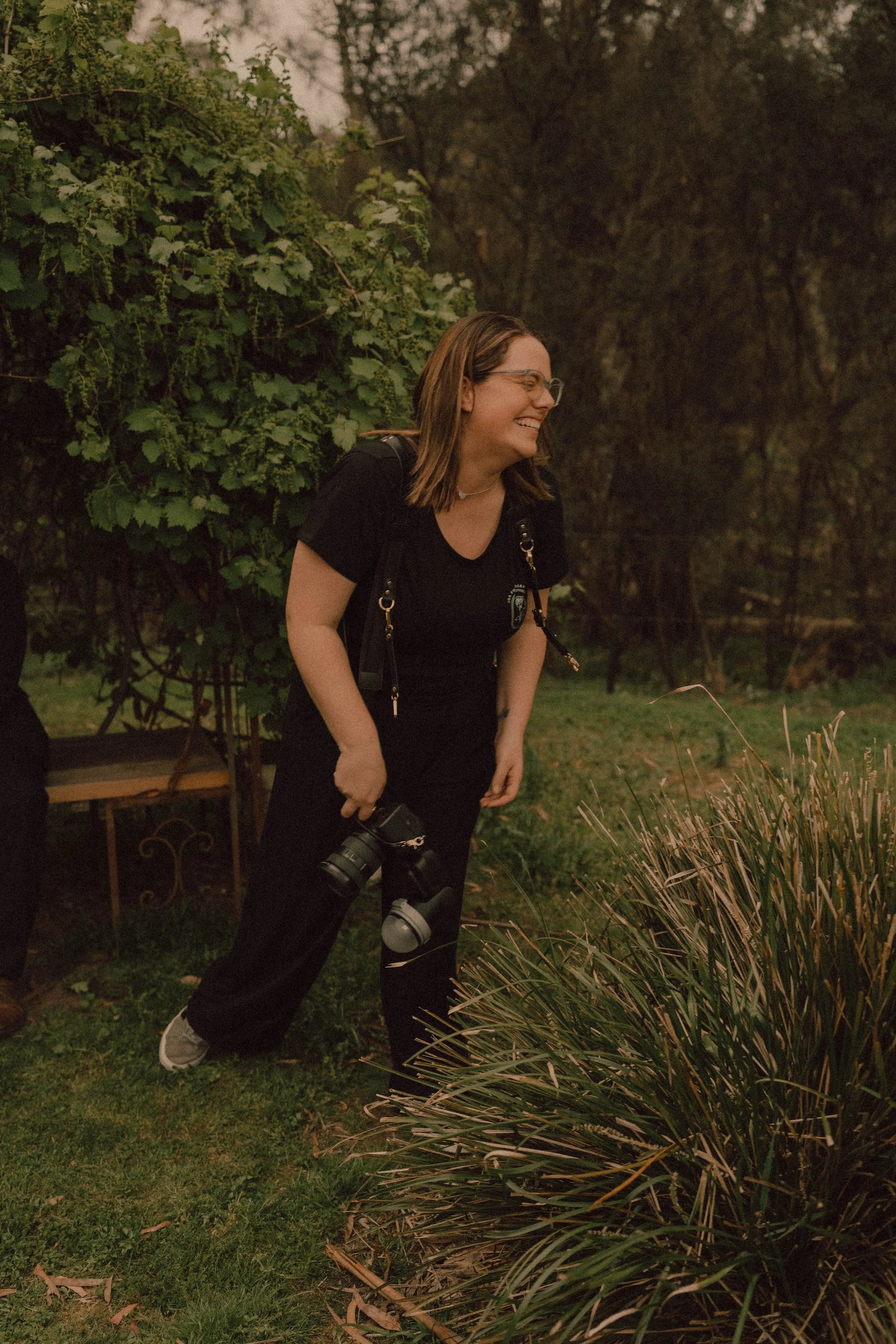 A photographer with glasses, wearing a black ada photography branded T-shirt and carrying a camera, is smiling and laughing forward in a garden with greenery and plants at bridges restaurant also known as woodland estate in hurstbridge