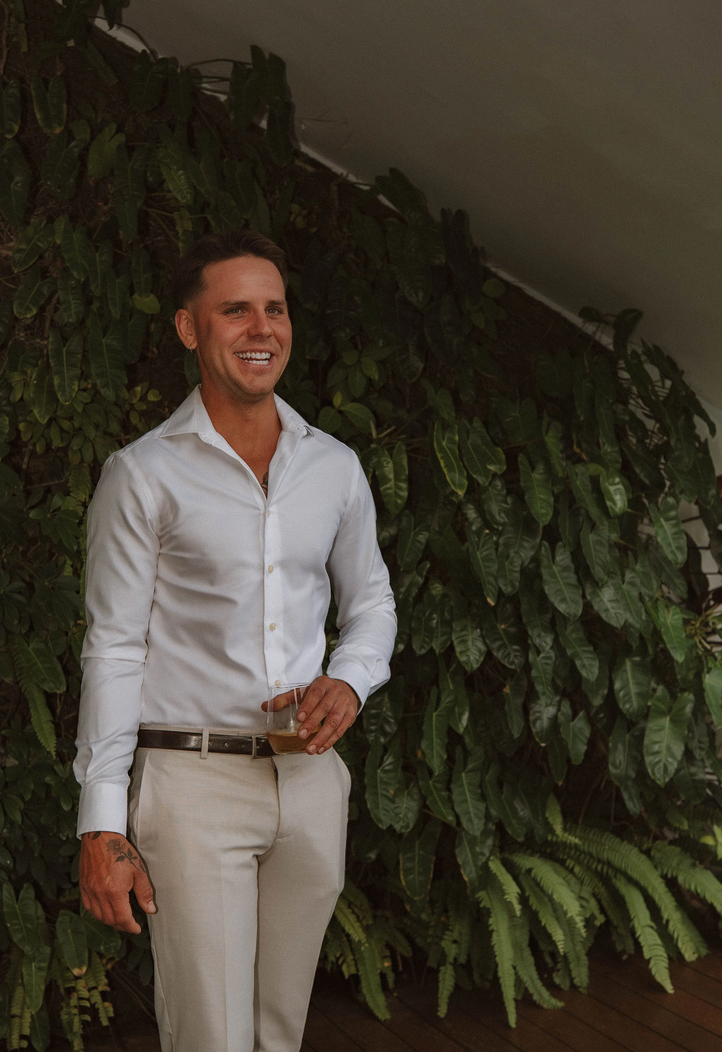 A smiling man in a white dress shirt and beige pants holding a drink in front of a wall of green plants at an indoor event. yarra valley and Melbourne wedding photographer