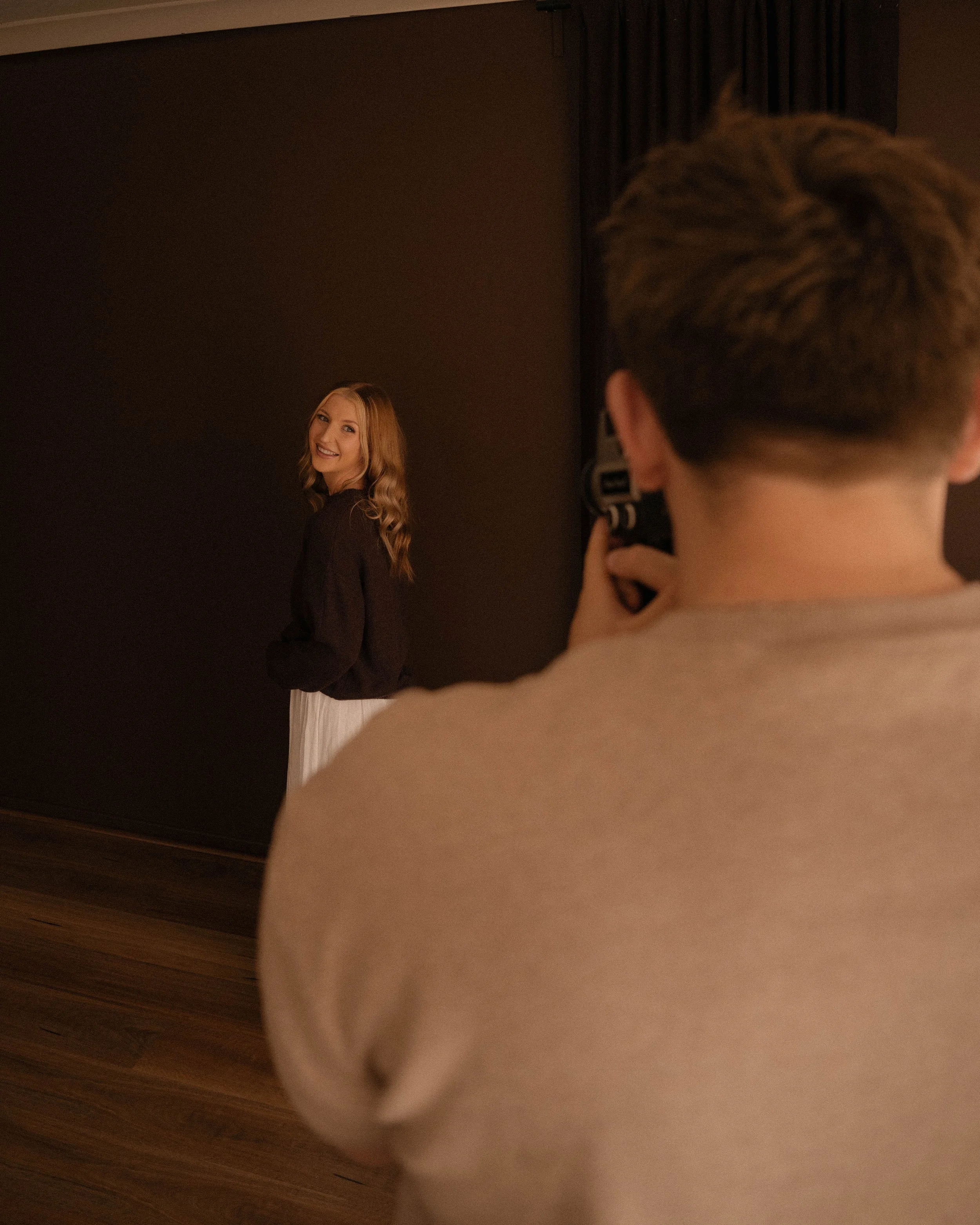 couples photoshoot in whittlesea studio, blonde women in a white dress and brown jumper smiling looking at her fiancé who is taking a photo of her, filming her laugh with a digital super 8