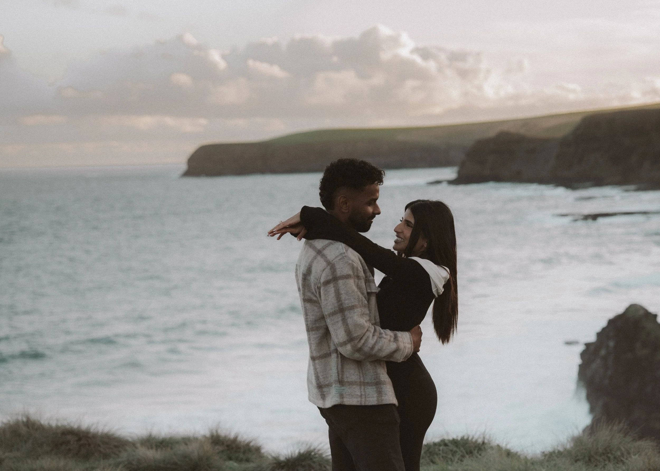 A couple embracing on a grassy shoreline with cliffs and ocean in the background, during sunset. moments after their proposal/engagement with jess a yarra valley and Melbourne wedding photographer